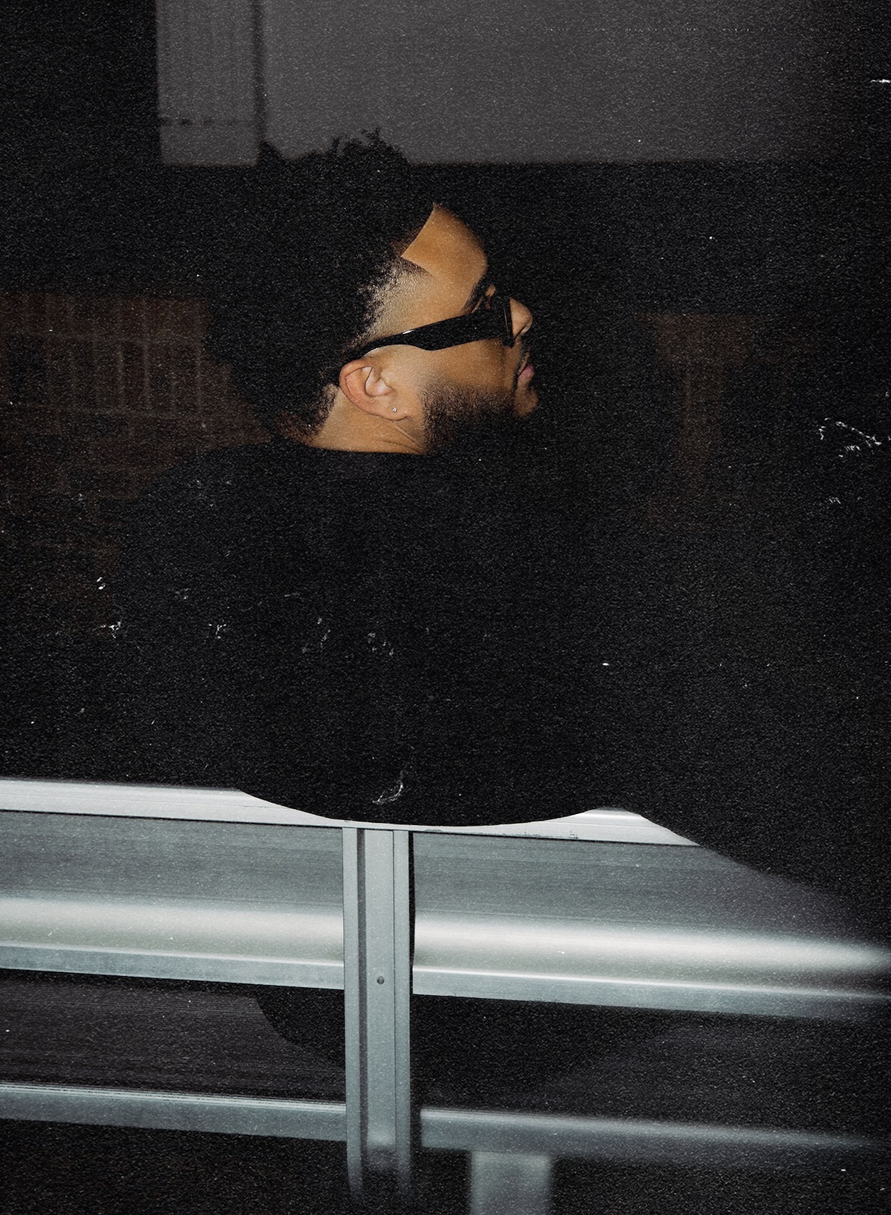 Person with glasses and curly hair sitting on a bench at night, facing sideways, in a dark environment.