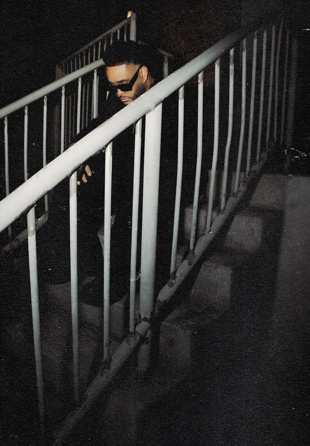 Person wearing sunglasses and dark clothing behind metal railing in dark environment.