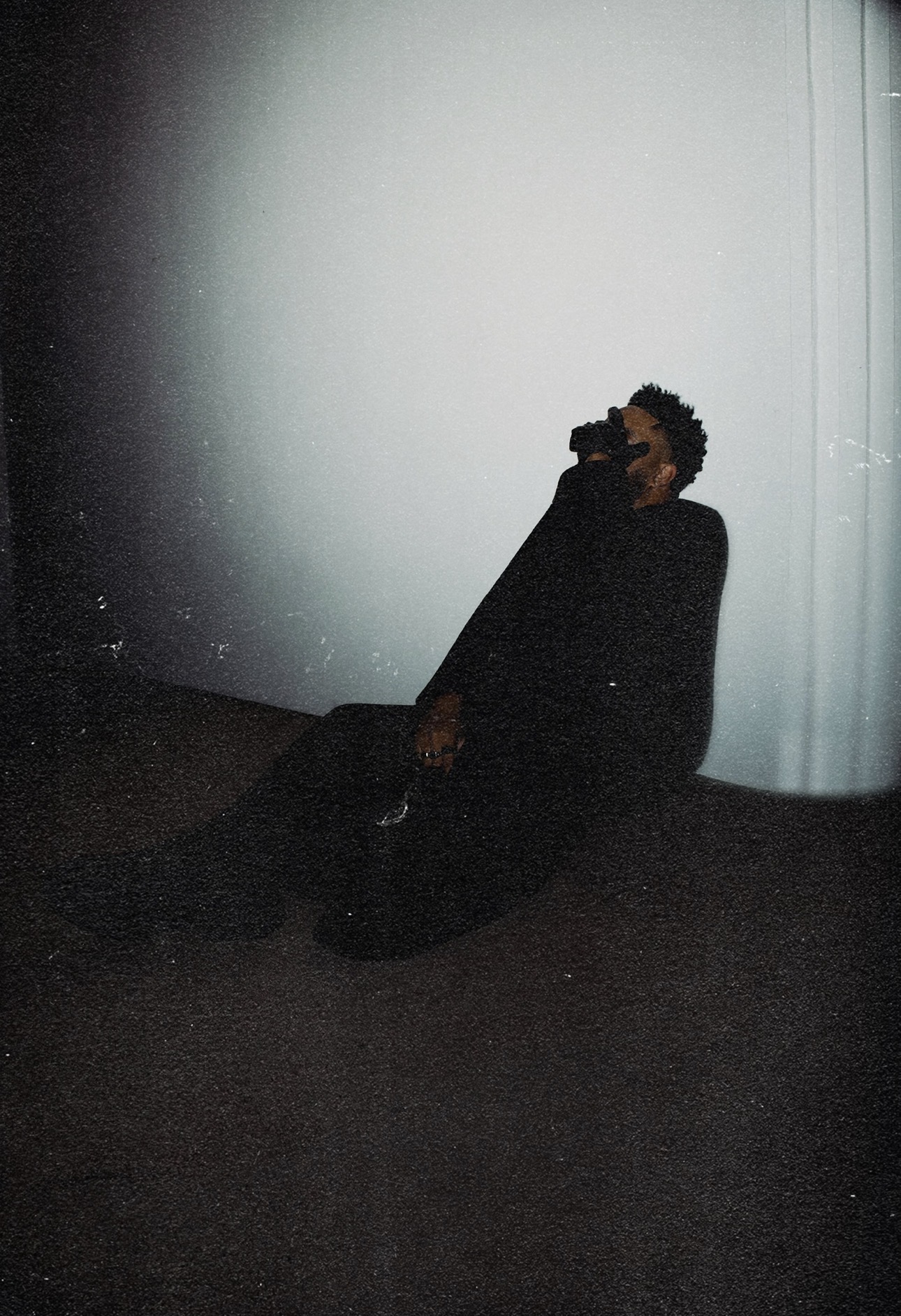 Person sitting on the floor against a light-colored wall, holding a camera to their face, in a dark room.