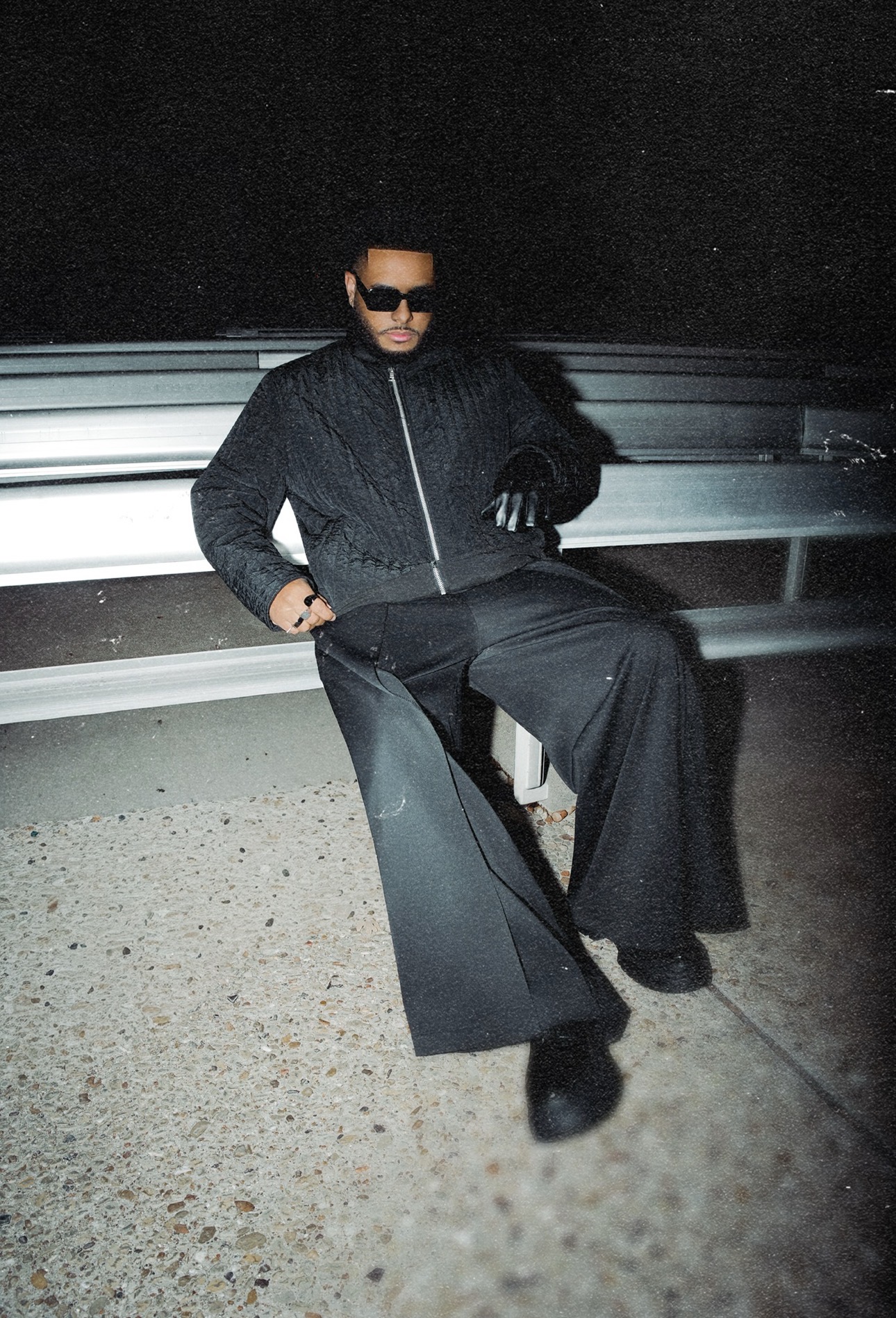 Person sitting on a guardrail at night, wearing sunglasses, a dark jacket, and wide-leg pants.