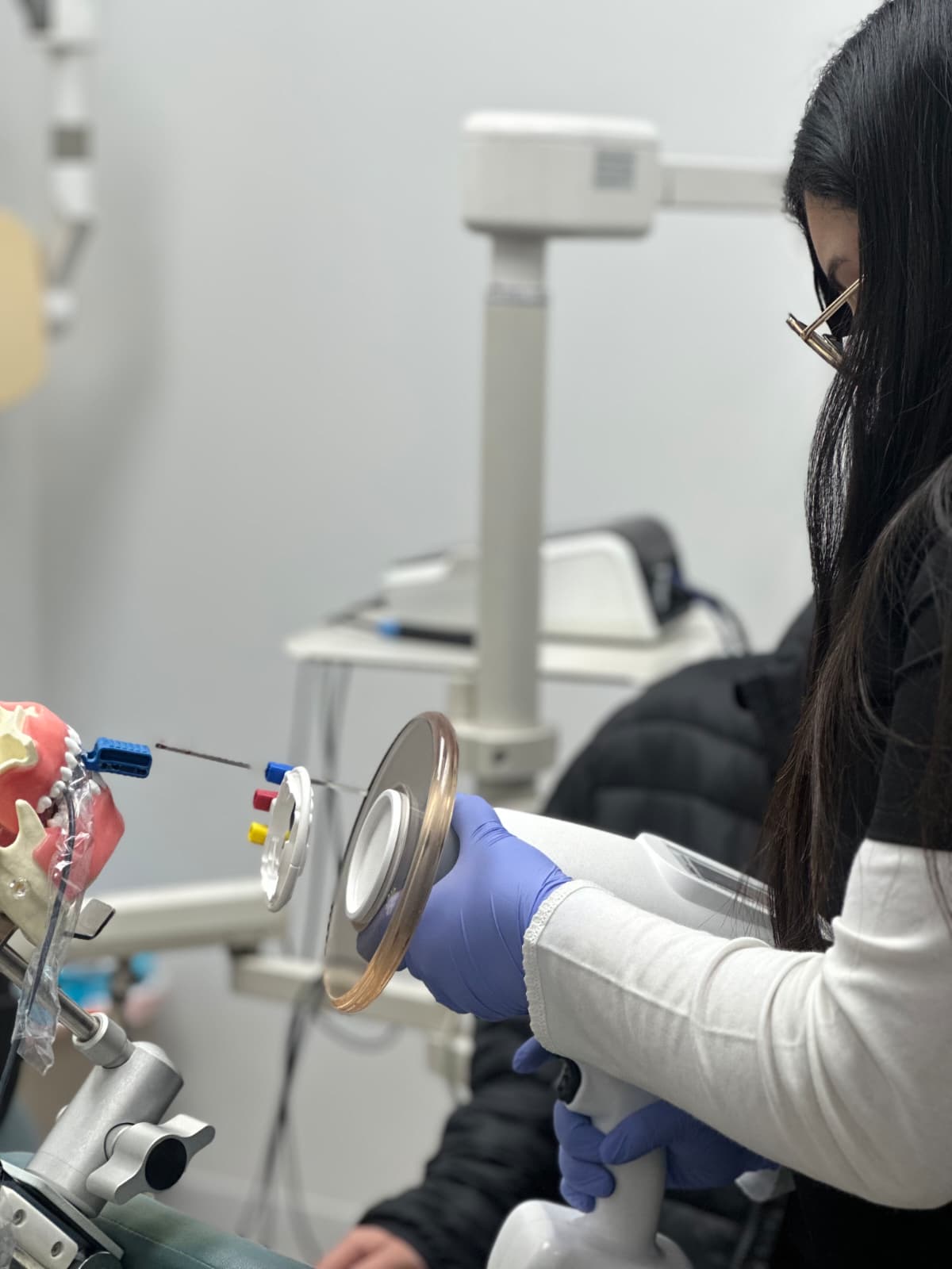 Person wearing gloves holding a dental tool in a clinical setting with dental equipment nearby.