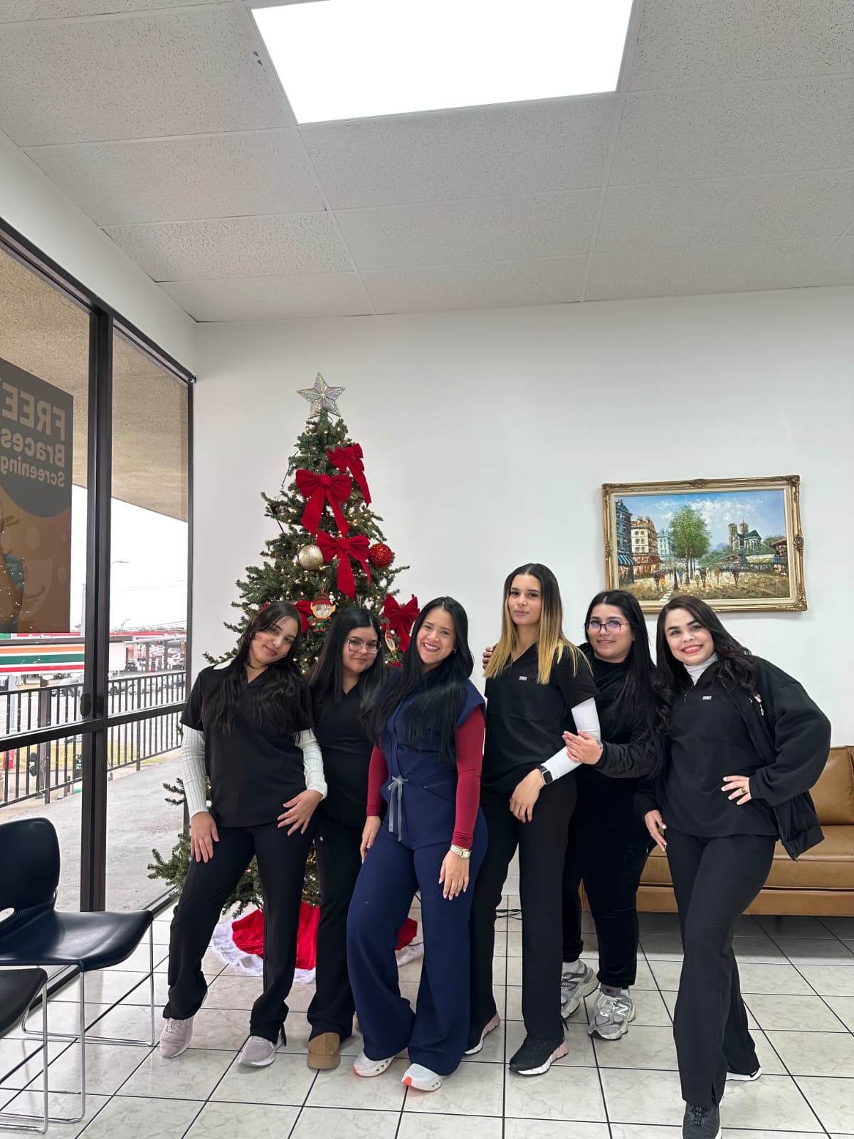 Five women standing indoors near a decorated Christmas tree, with a window and framed painting behind them.