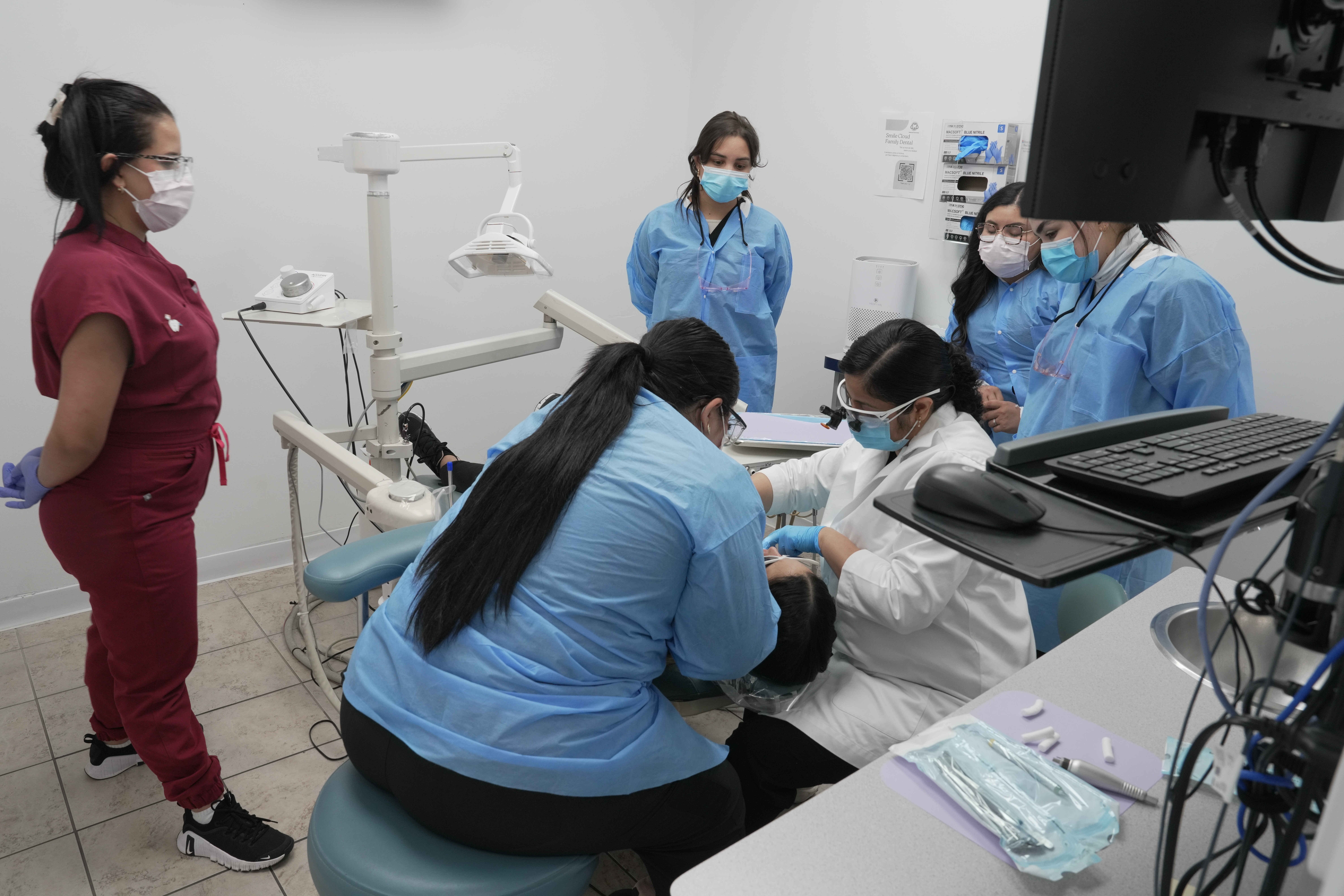 Dental team in protective gear attending to patient in clinic, with staff observing, monitors, and dental equipment visible.