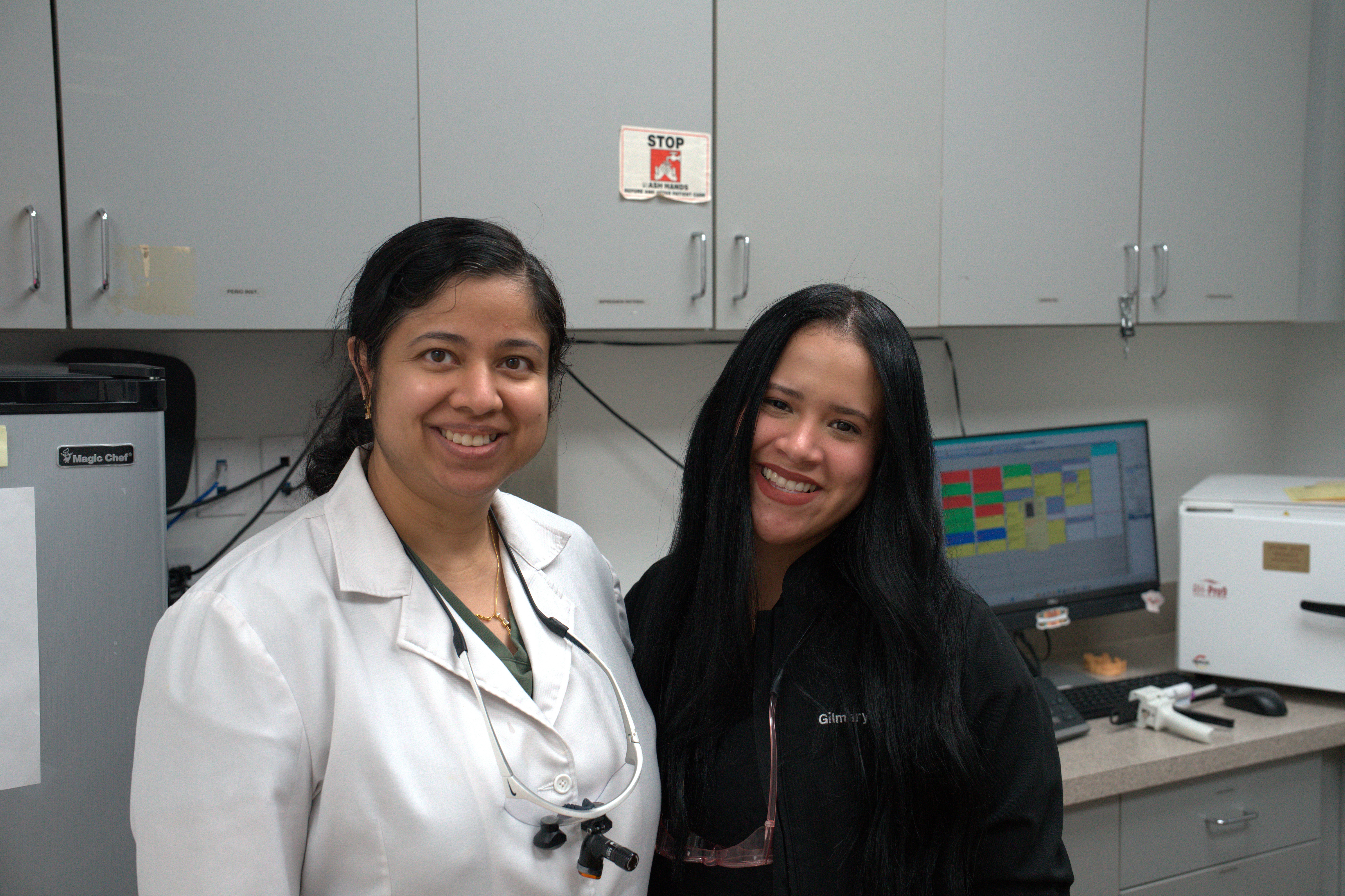 Two women in lab coats smiling in a laboratory with equipment and cabinets behind them.