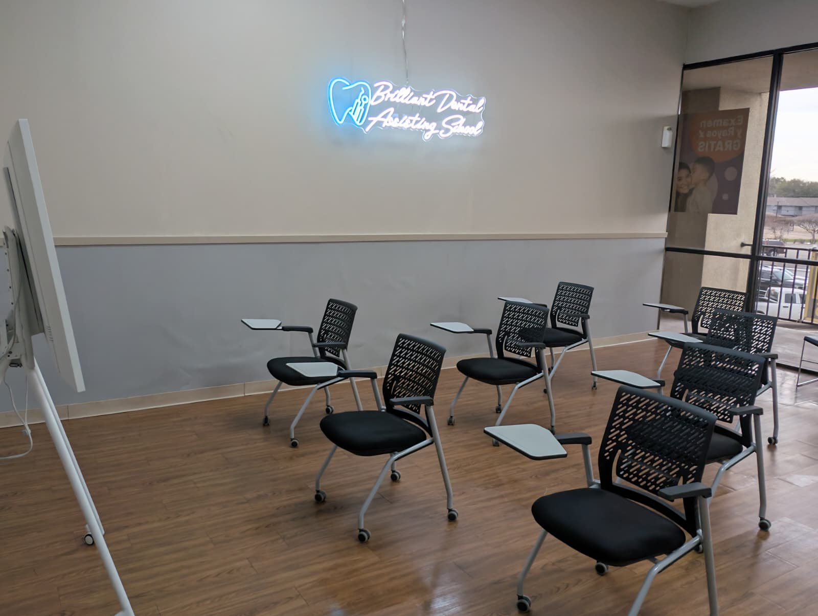 Empty classroom with chairs and desks, neon sign on wall, large window showing outdoor view.