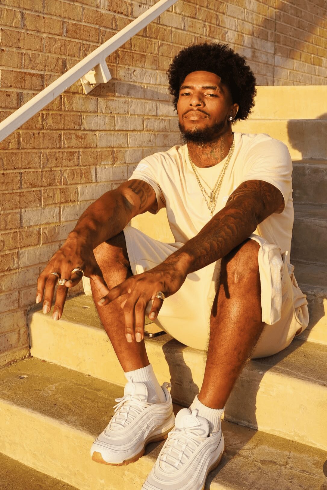 Young man with curly hair and beard sitting on stairs outdoors, wearing white t-shirt, shorts, and sneakers.