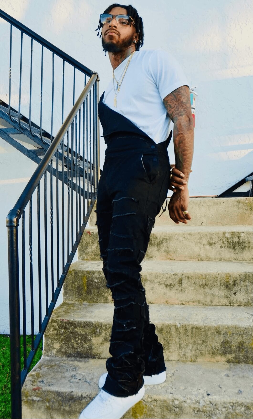 Man with sunglasses and tattoos standing on stairs, leaning on railing outdoors, wearing a light blue t-shirt and black pants.