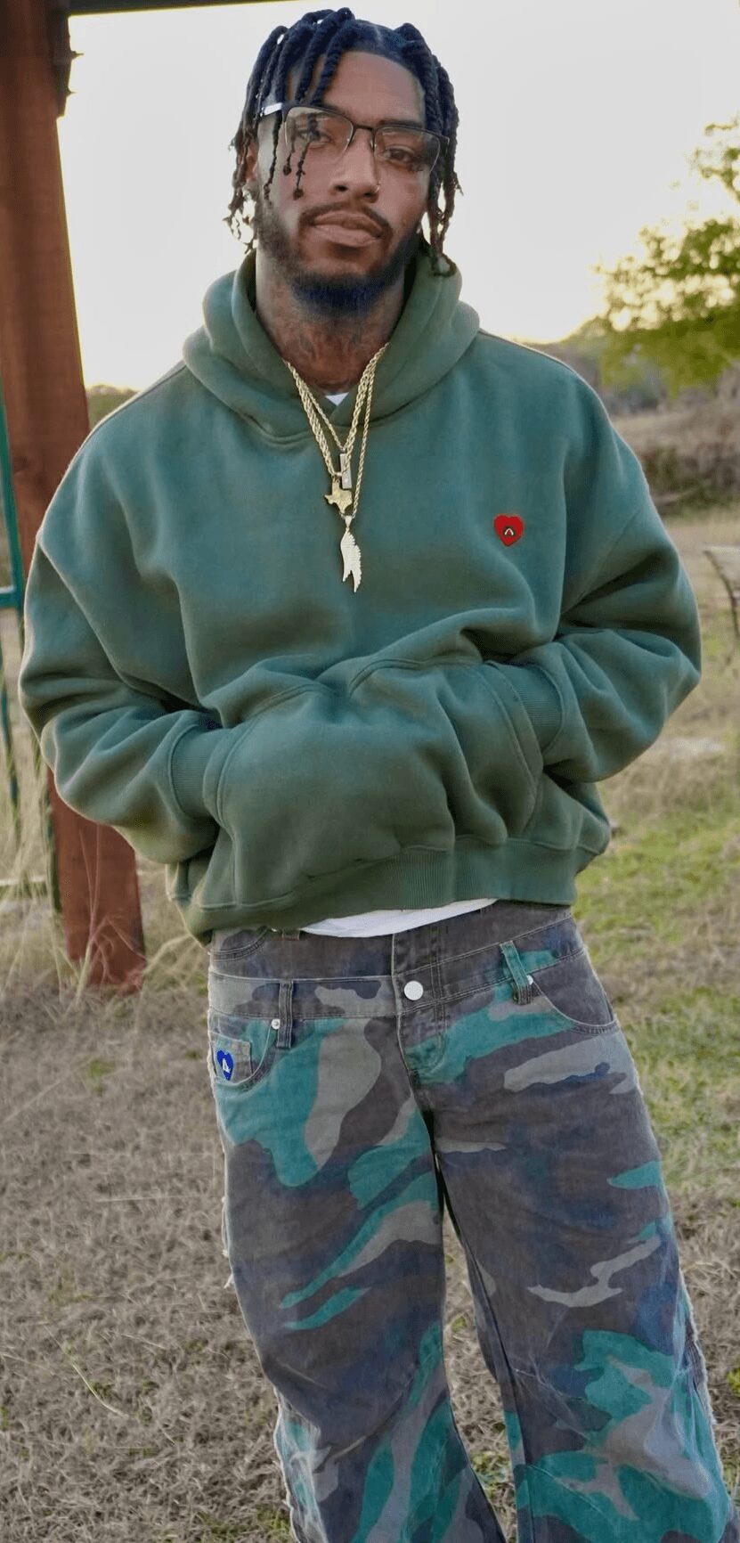 Young man with dreadlocks wearing a green hoodie and camouflage pants outdoors.