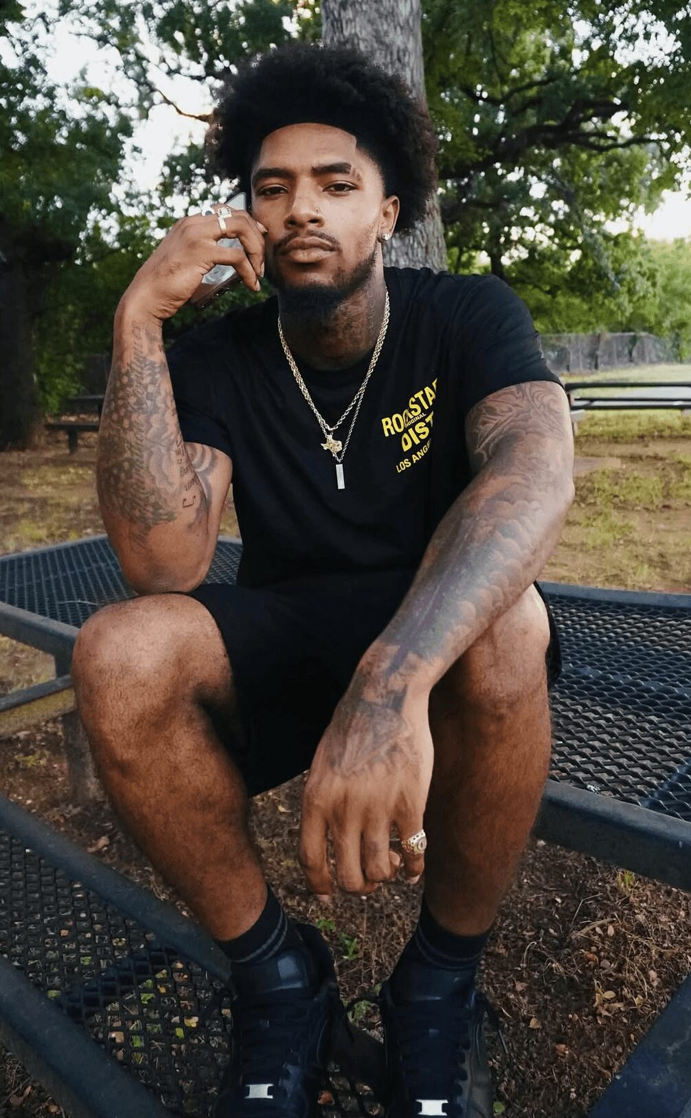 Young man sitting on a park bench with trees in background, wearing black t-shirt and shorts, looking at camera.