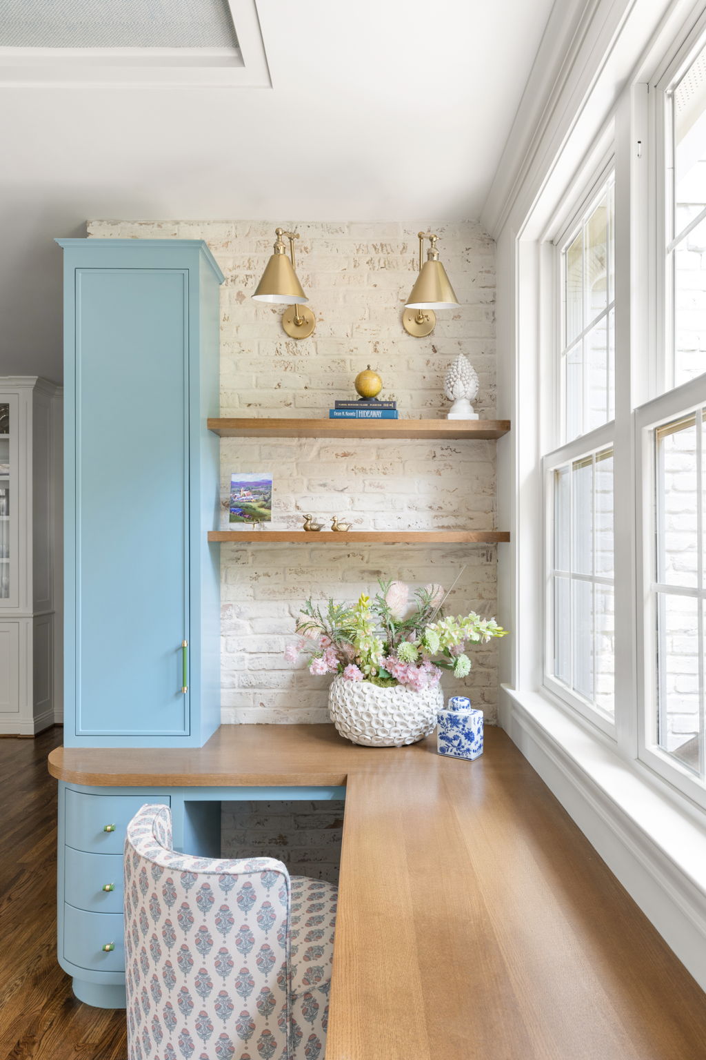 Corner of a room with a blue cabinet, two wall-mounted lamps, floating shelves, a basket of flowers, and large windows.