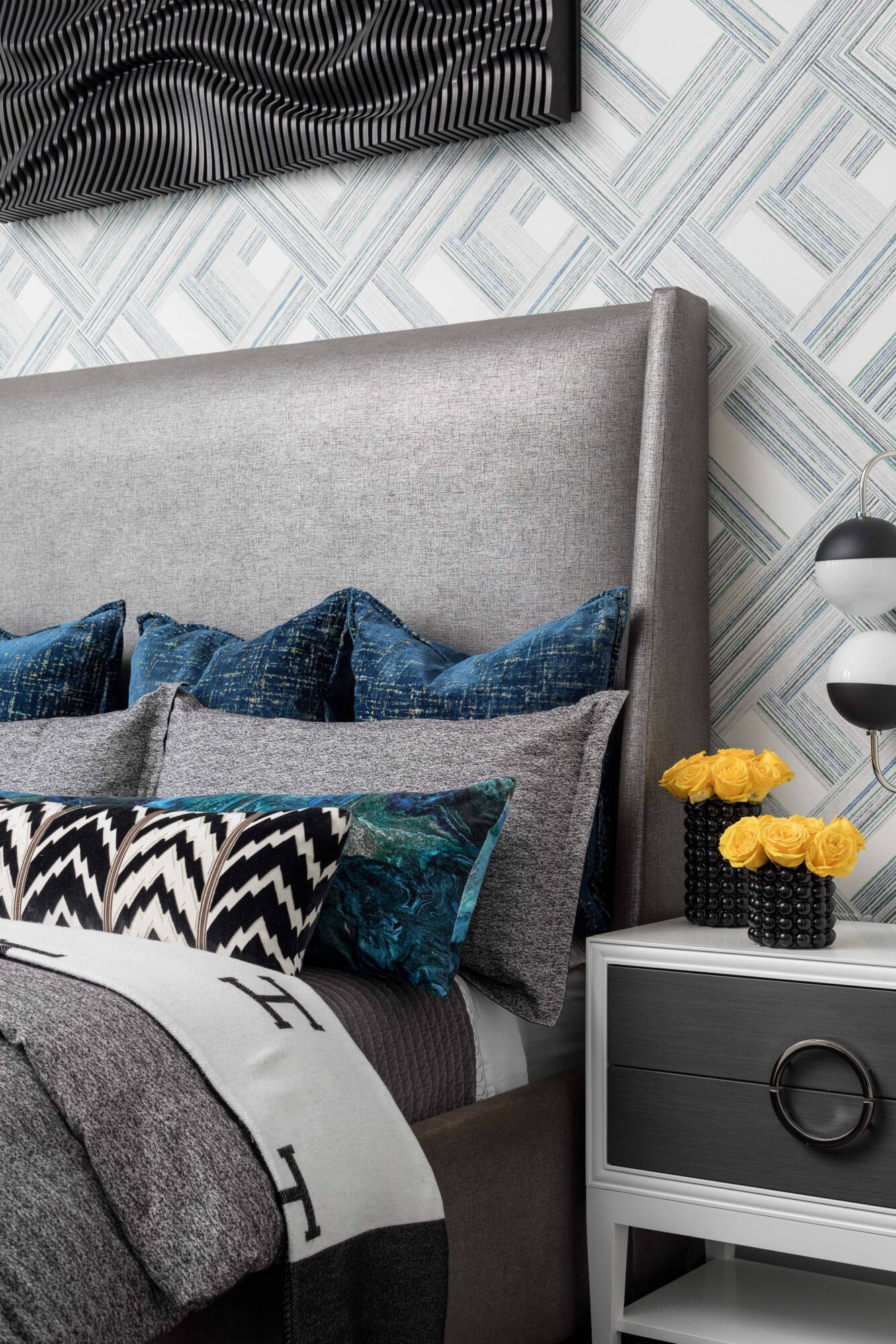 Close-up of a bedroom with a gray upholstered headboard, patterned pillows, and a white nightstand with yellow flowers.