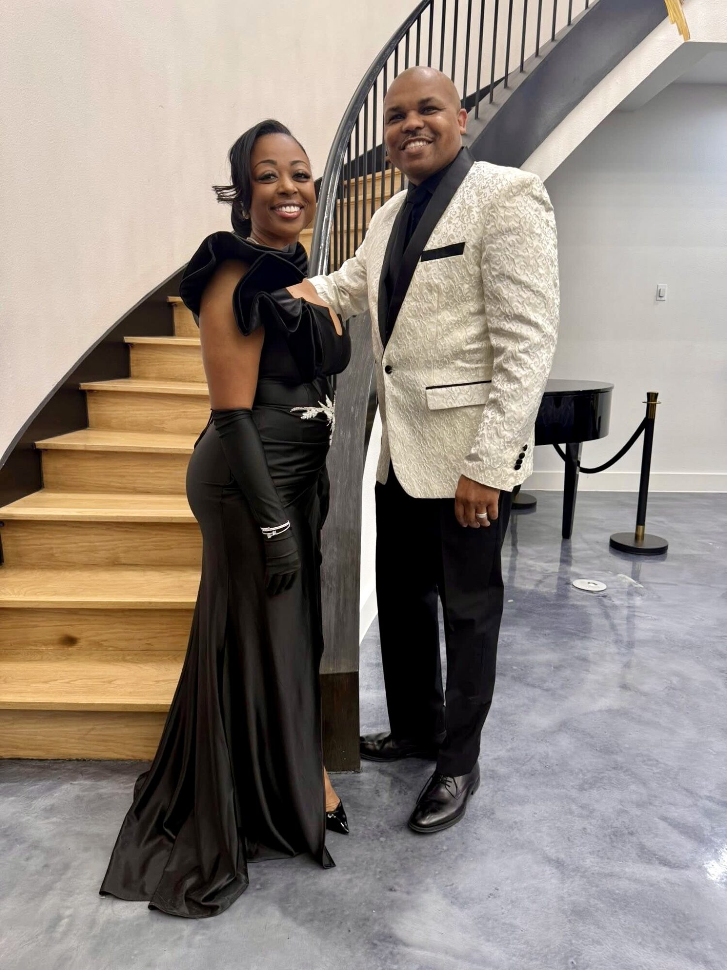 A woman and man pose together indoors, smiling, with a staircase in the background. The woman wears a black gown, the man a white blazer.