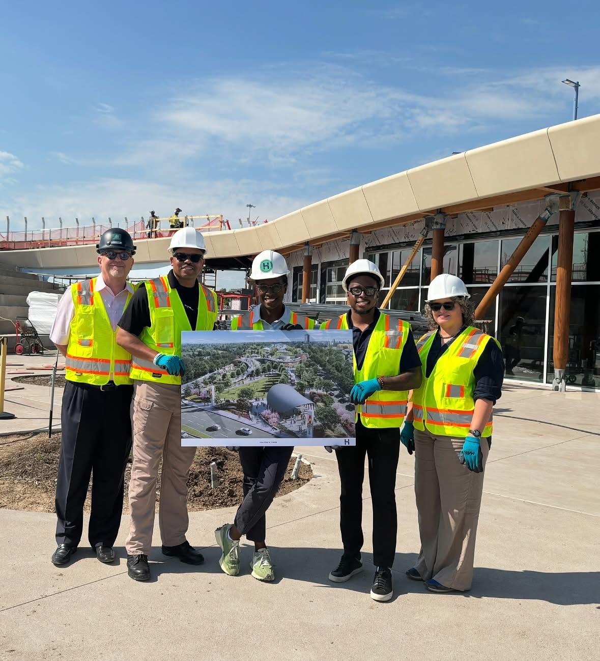 Five people in safety vests and helmets holding a large map or plan outdoors near a construction site.
