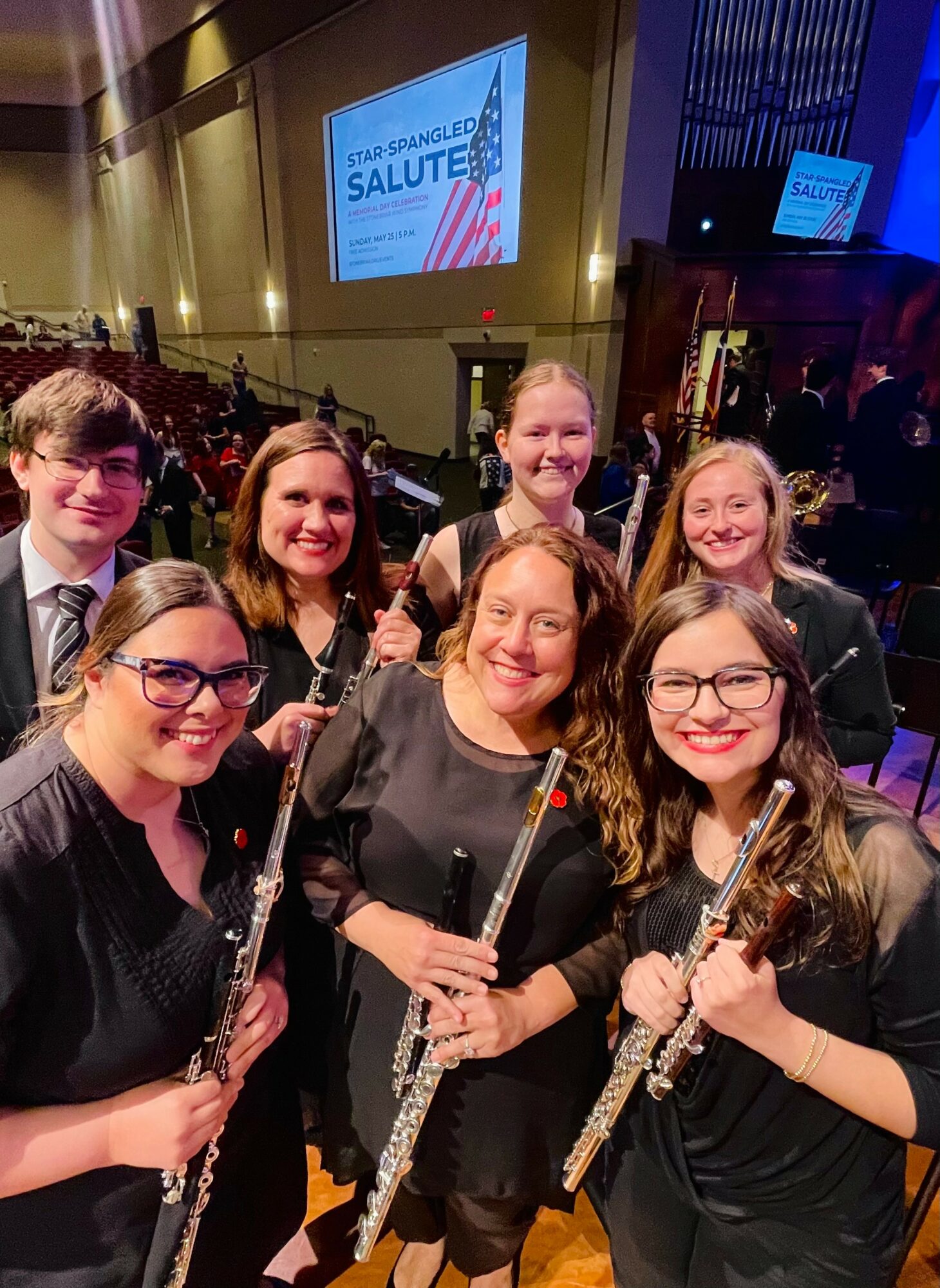 Group of seven women holding flutes at an indoor event with a stage and screen in background.