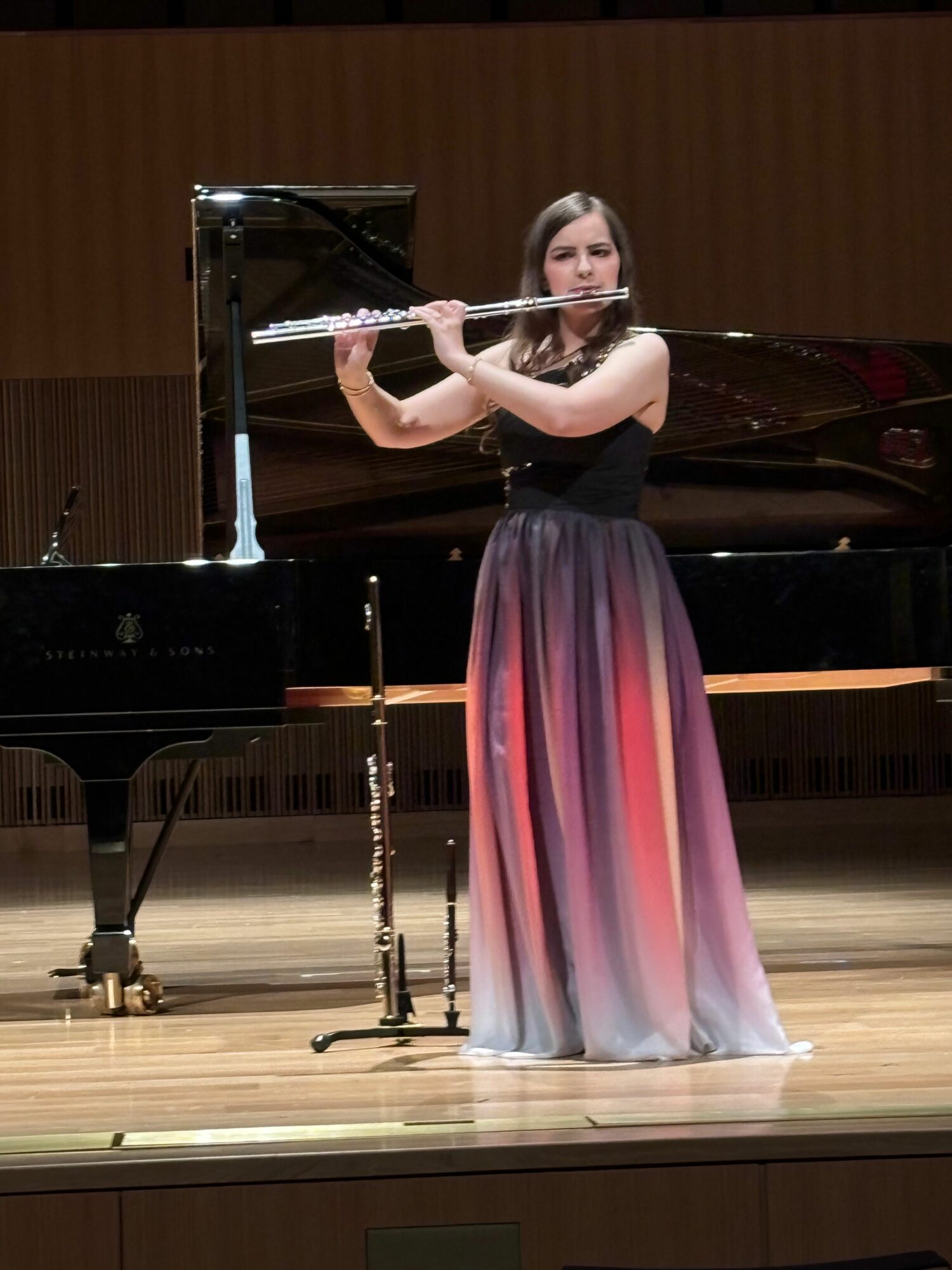 Young woman playing flute on stage with grand piano behind her, wearing a long gradient dress, wooden floor, wooden panel background.