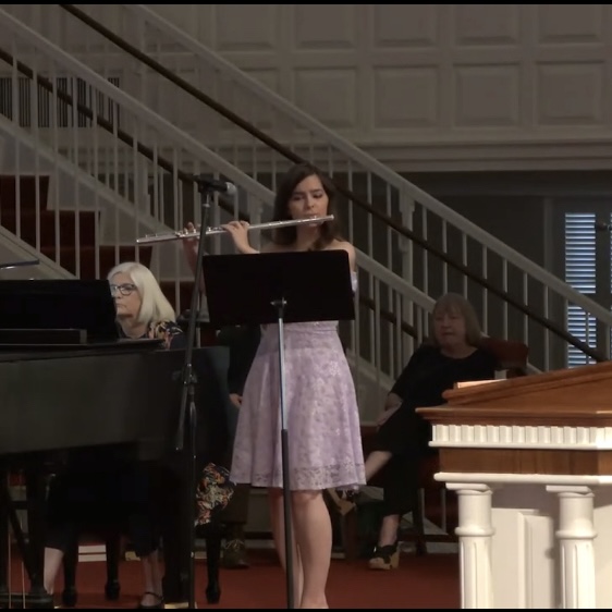 Young woman playing flute at microphone, with two seated women and piano in background, in a formal indoor setting.