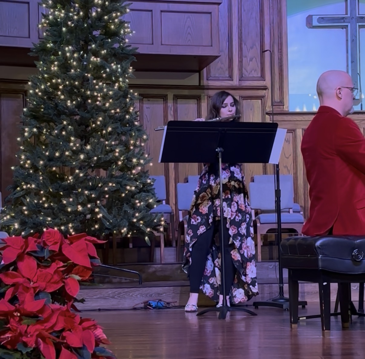 Woman playing a music stand near a decorated Christmas tree, with a man in red nearby, in a wooden-paneled room.