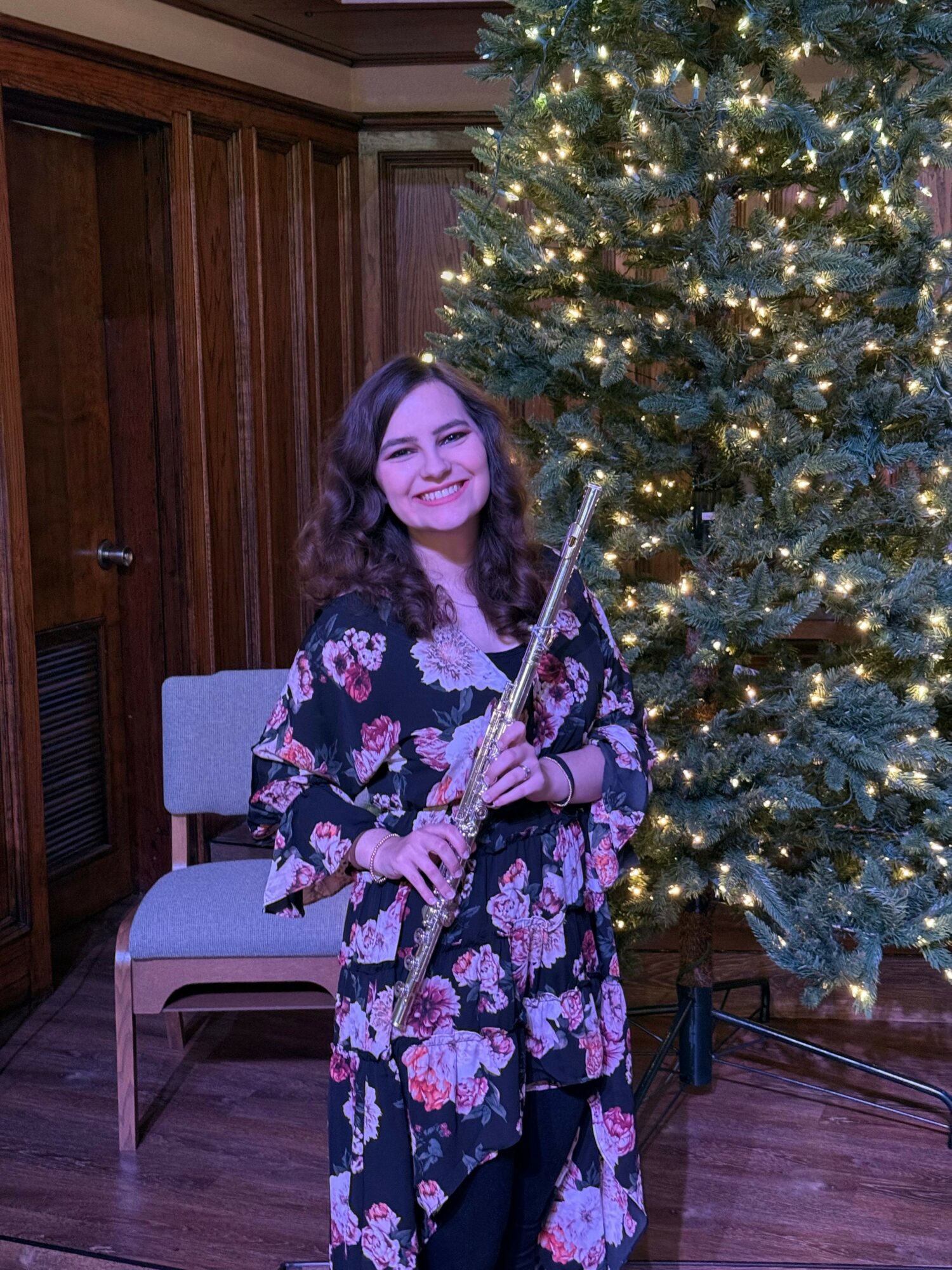 Young woman with dark curly hair holding a flute, standing next to a decorated Christmas tree with lights.