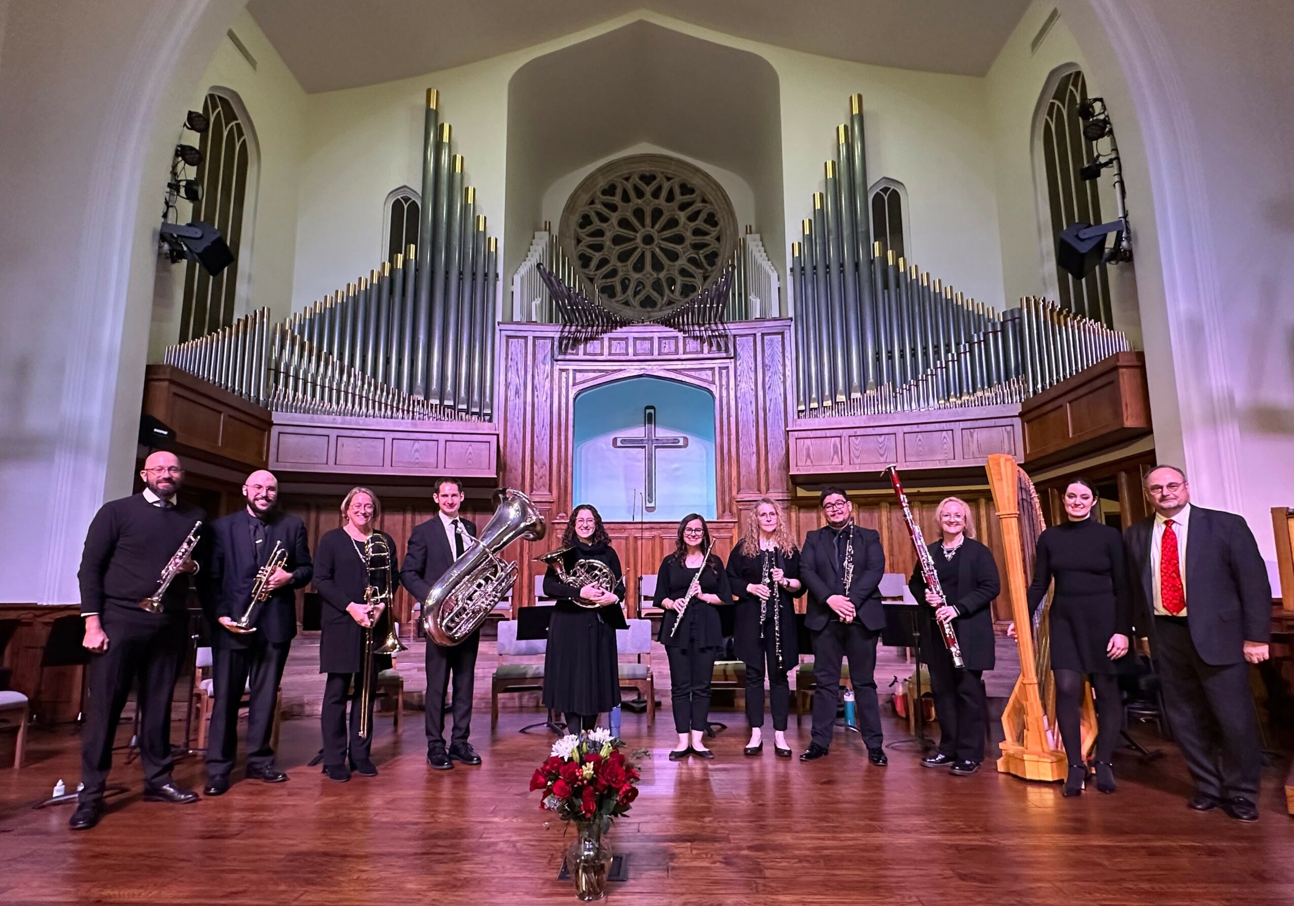 Group of musicians standing on stage in front of large pipe organ inside church, with a cross and floral arrangement in front.