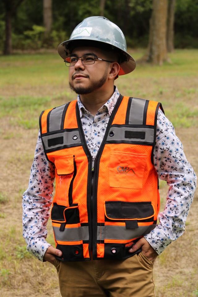 Young man wearing safety helmet and reflective vest outdoors with trees and grass in background.