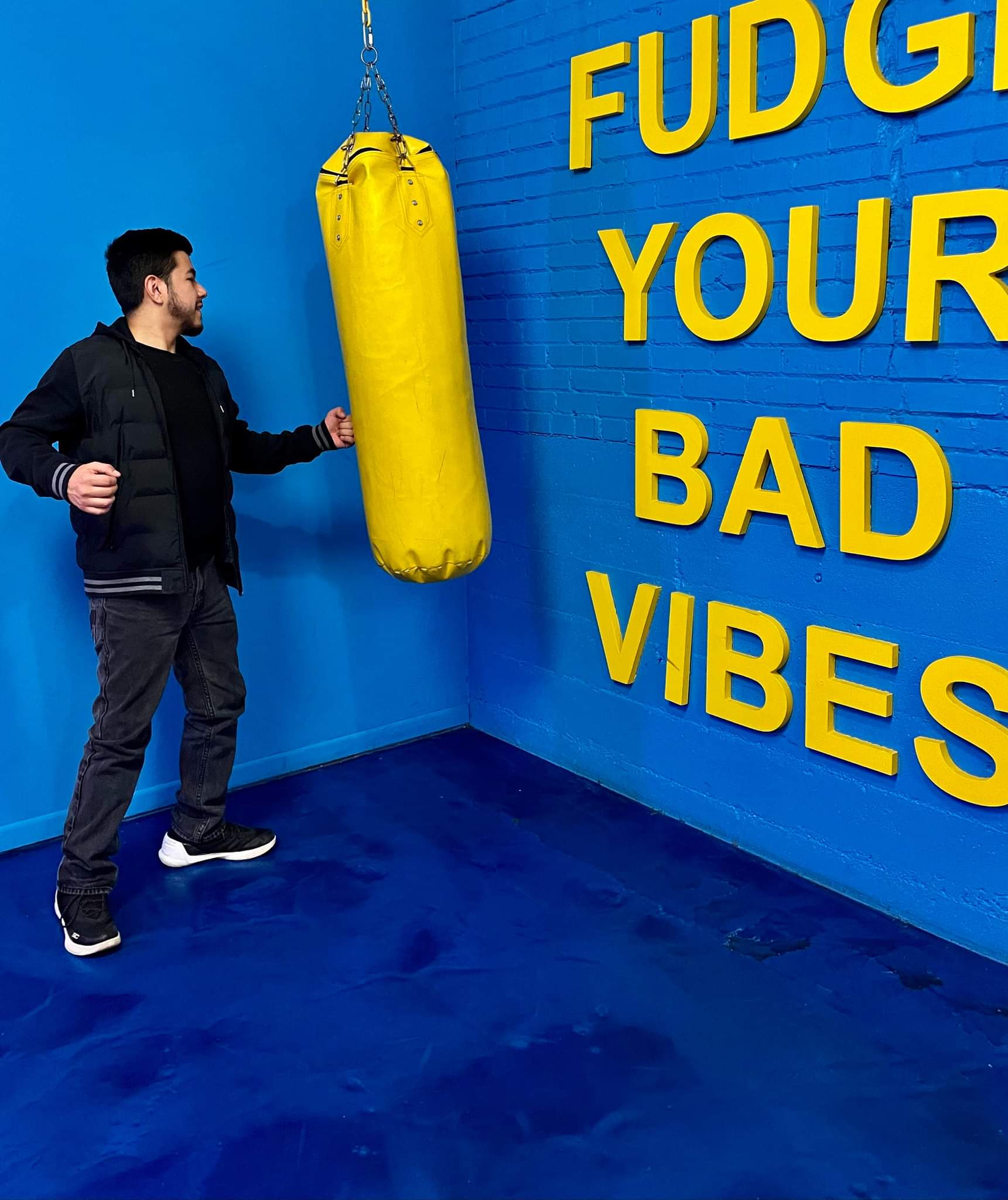Man in black jacket and jeans standing near yellow punching bag in blue room with yellow text on wall.