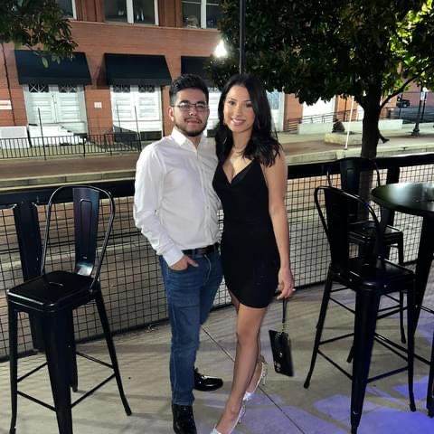 A man and woman stand together outdoors at night, smiling, with empty chairs and a fence behind them.