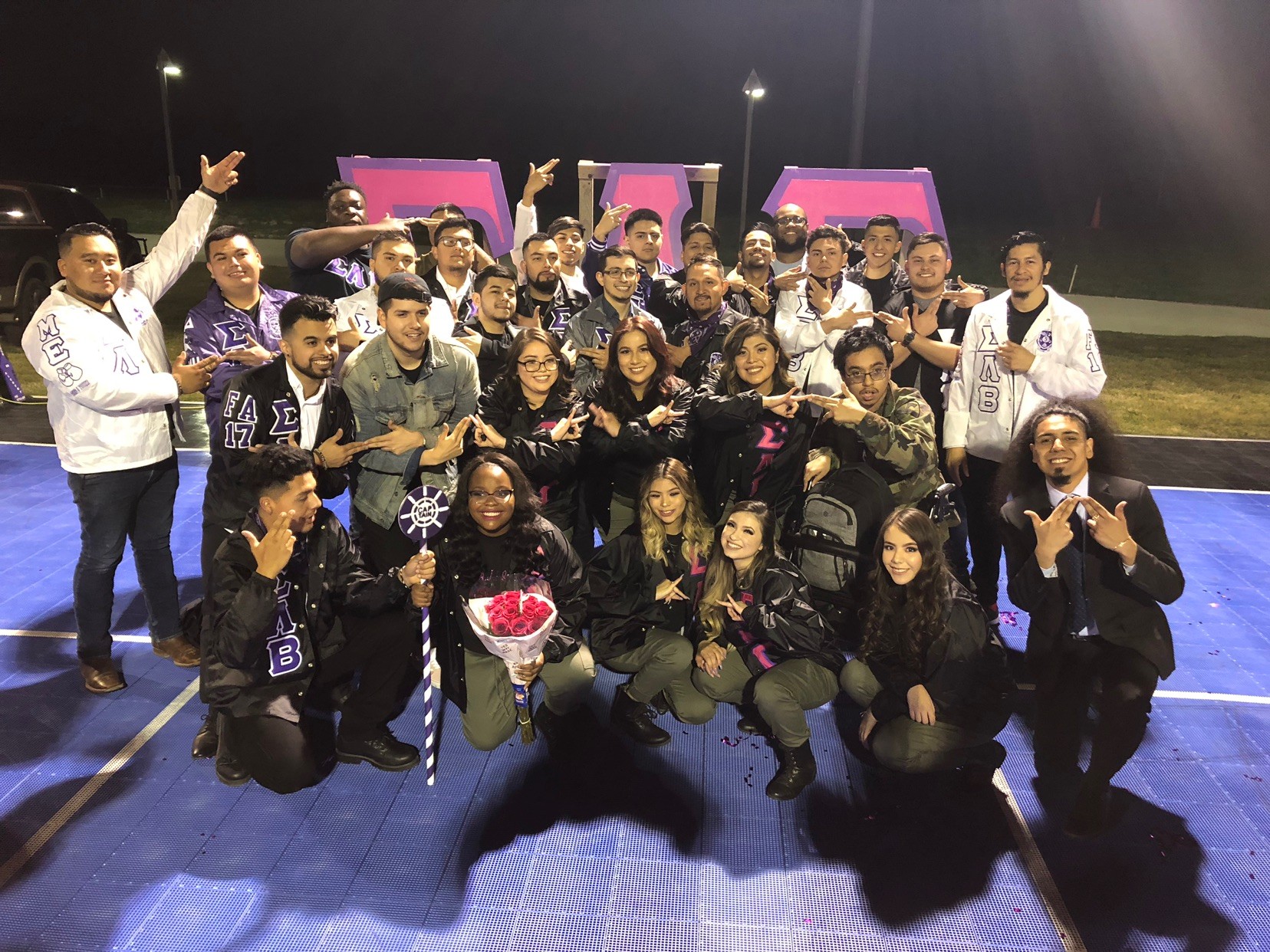 Group of people gathered outdoors on a blue surface, some holding flowers, posing for a photo at night.