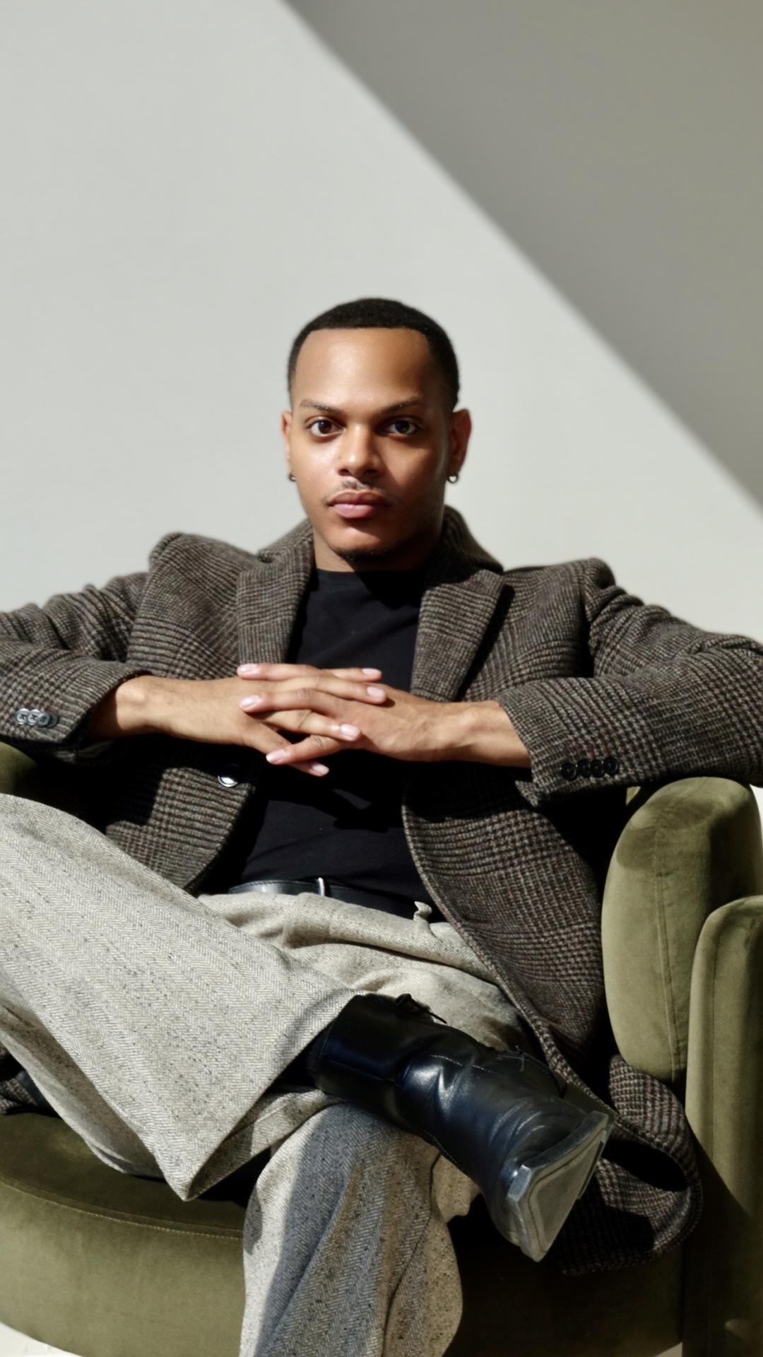Person sitting on a green chair with crossed legs, wearing a blazer and black shoes, against a plain background.