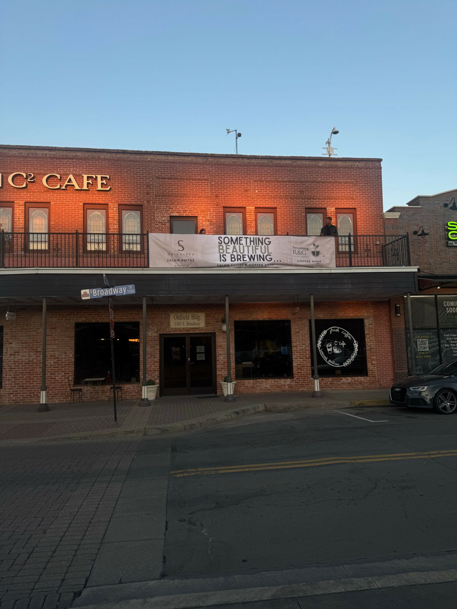 Two-story brick building with a sign reading 'SOMETHING IS BREWING' on the upper balcony, and a circular logo on the lower facade.