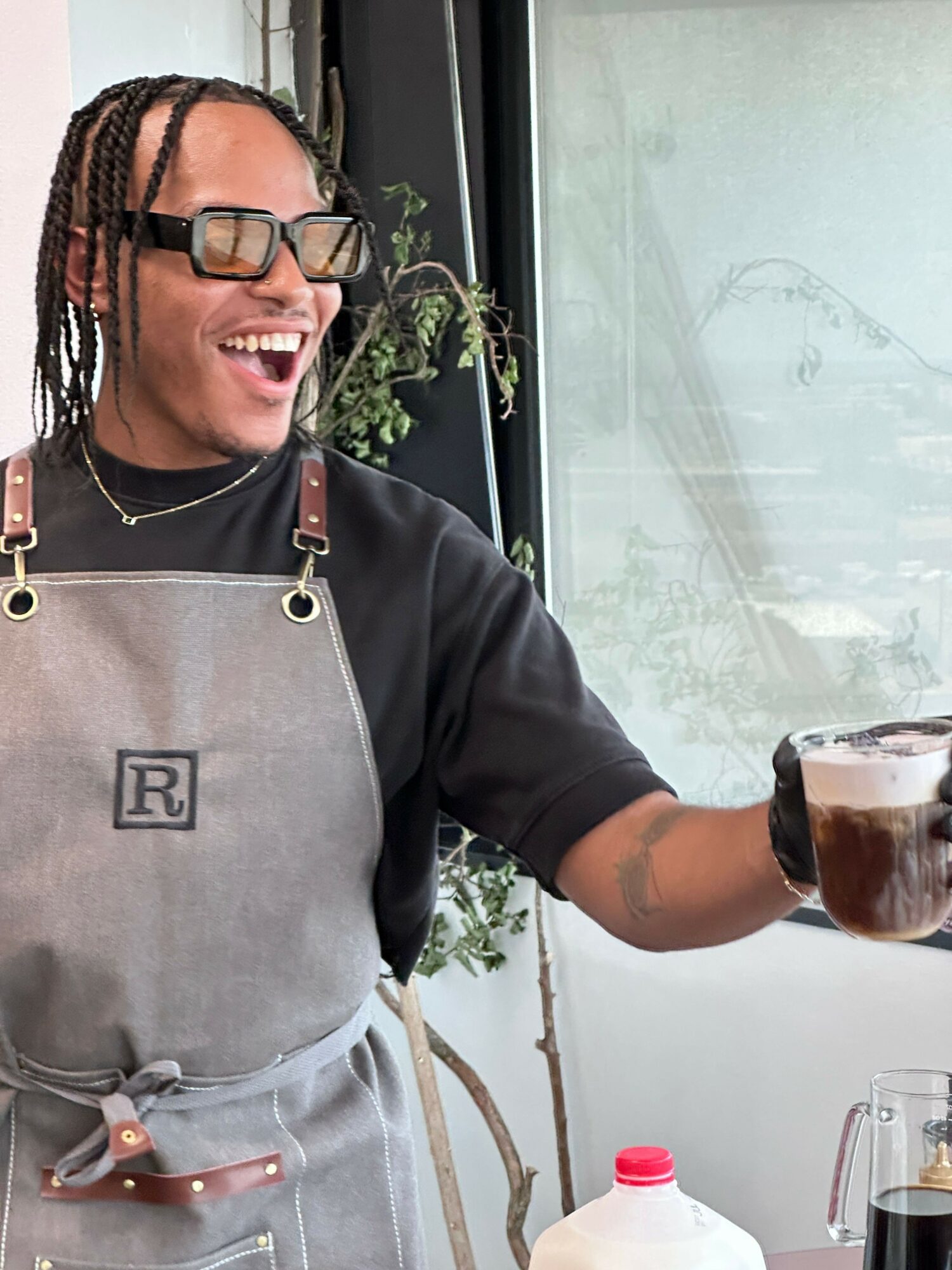 Person with glasses and braided hair smiling, holding a glass of dark beverage, wearing an apron, indoors near window.