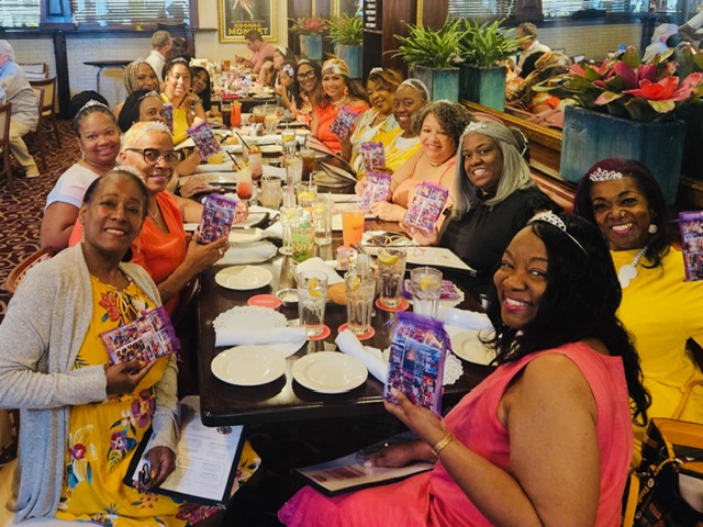 Group of diverse people sitting at a long dining table in a restaurant, smiling, holding gifts, with food and drinks on the table.