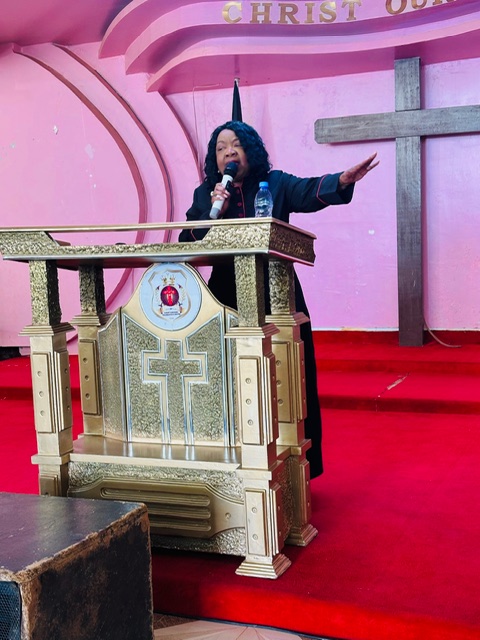 Woman speaking at a church podium with a cross and pink background, red carpet, and a large cross on the wall.