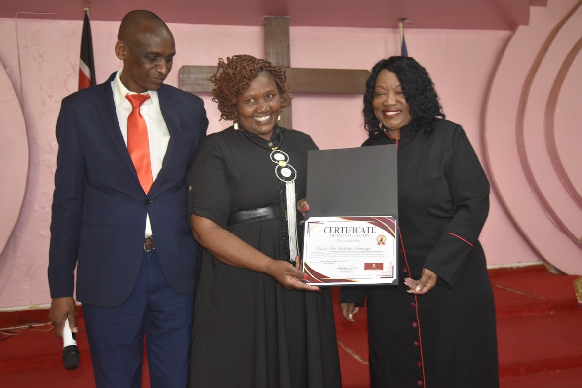 Three people standing on a stage, holding a certificate, smiling, with a pink background and red carpet.
