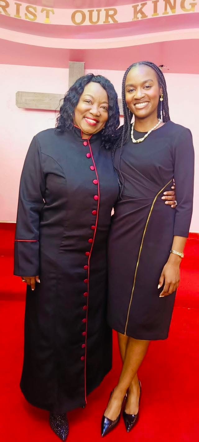Two women standing together, smiling, in formal dresses, on a red carpet with pink background and banner.
