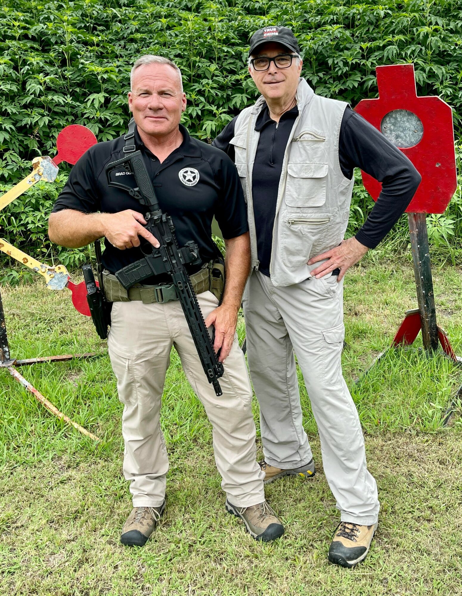 Two men standing outdoors, one holding a rifle, surrounded by greenery and target practice equipment.