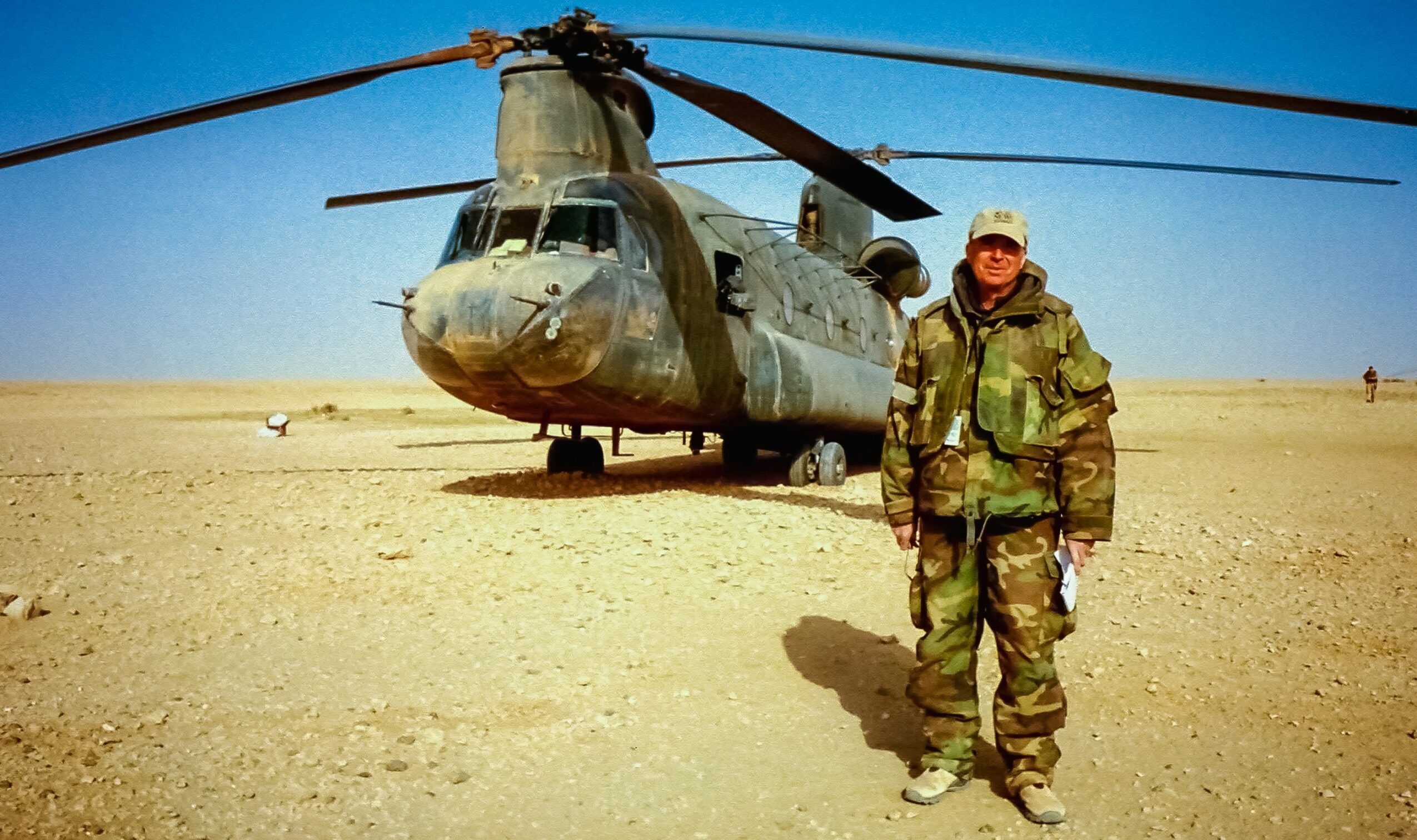 Soldier in camouflage uniform standing next to a military helicopter on desert terrain.