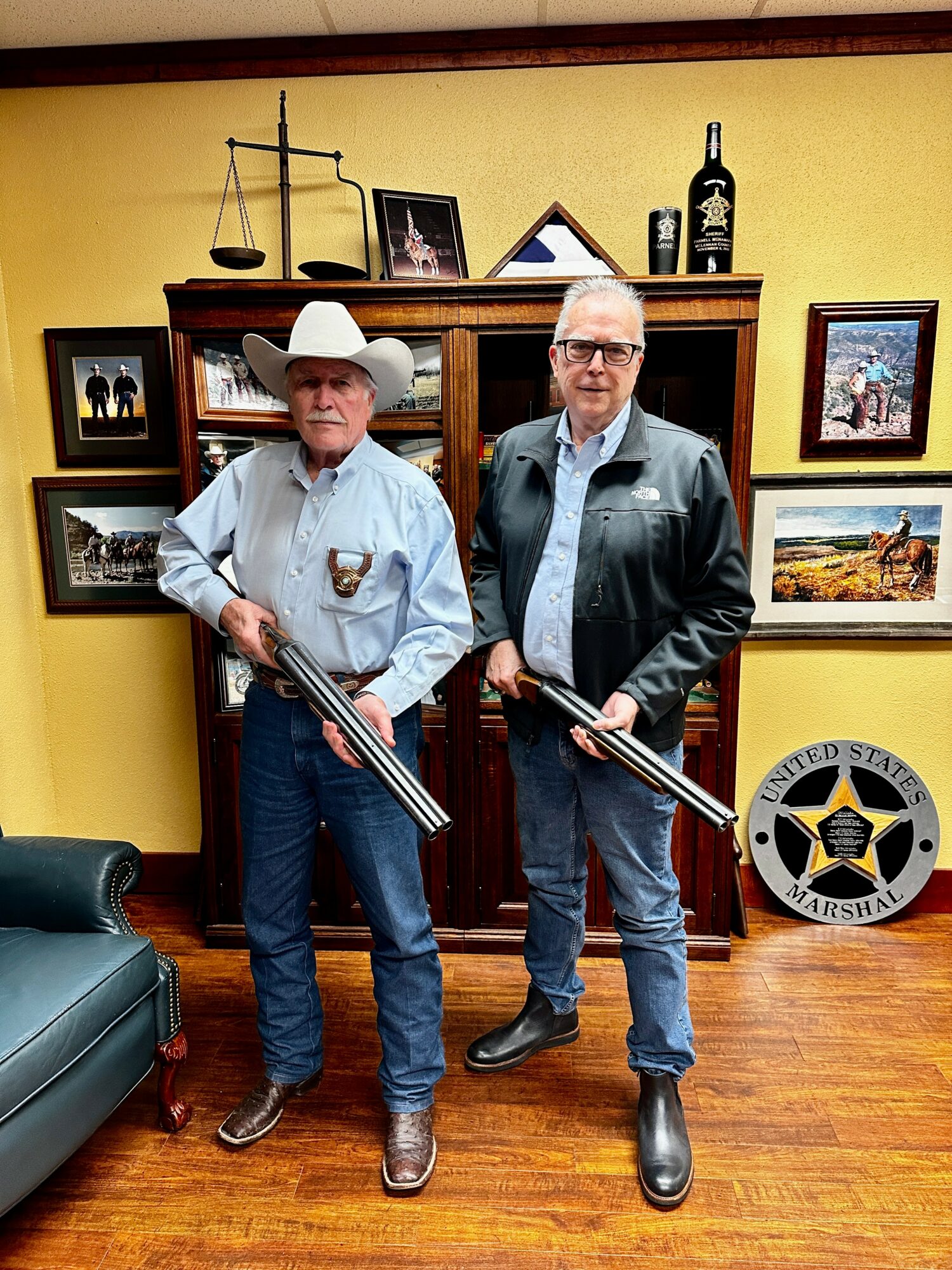 Two men holding rifles stand in a room with yellow walls, wooden furniture, and framed pictures.