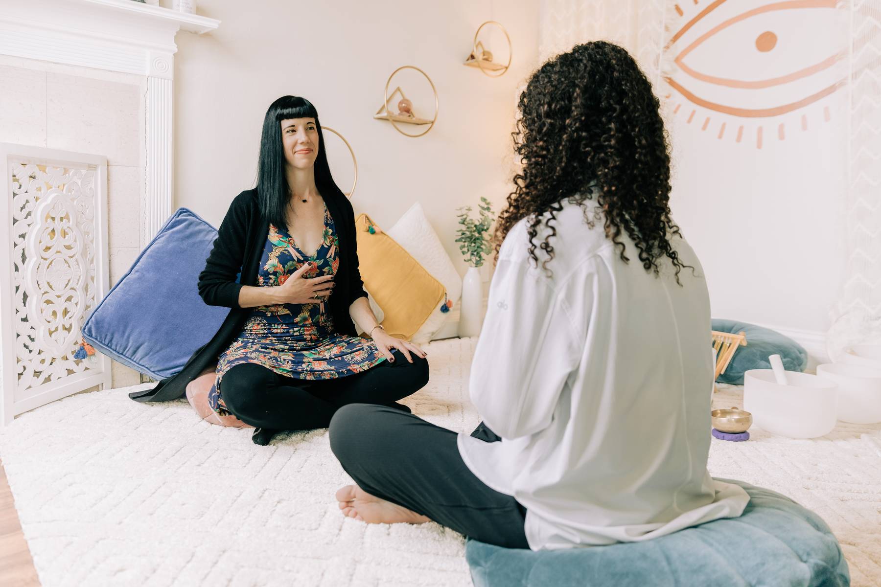 Two women sit on a white bed in a bright room, facing each other, engaged in conversation.
