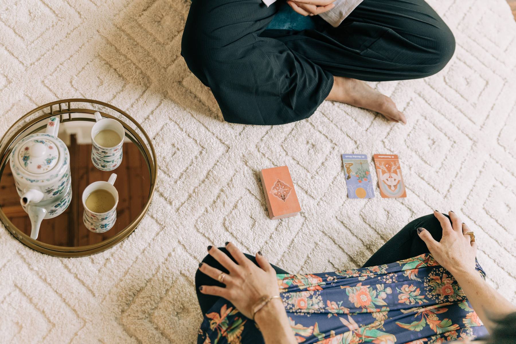 Person sitting cross-legged on a cream textured rug, with a tray of drinks and tarot cards nearby.