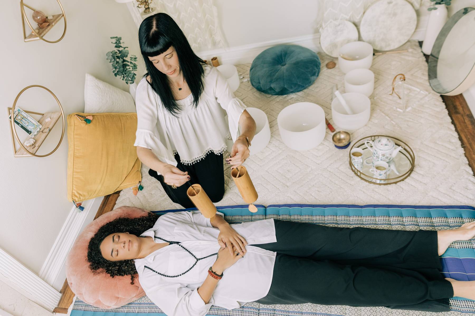 A woman lying on a pink pillow with a stethoscope, a woman standing nearby, in a cozy room with cushions and decor.
