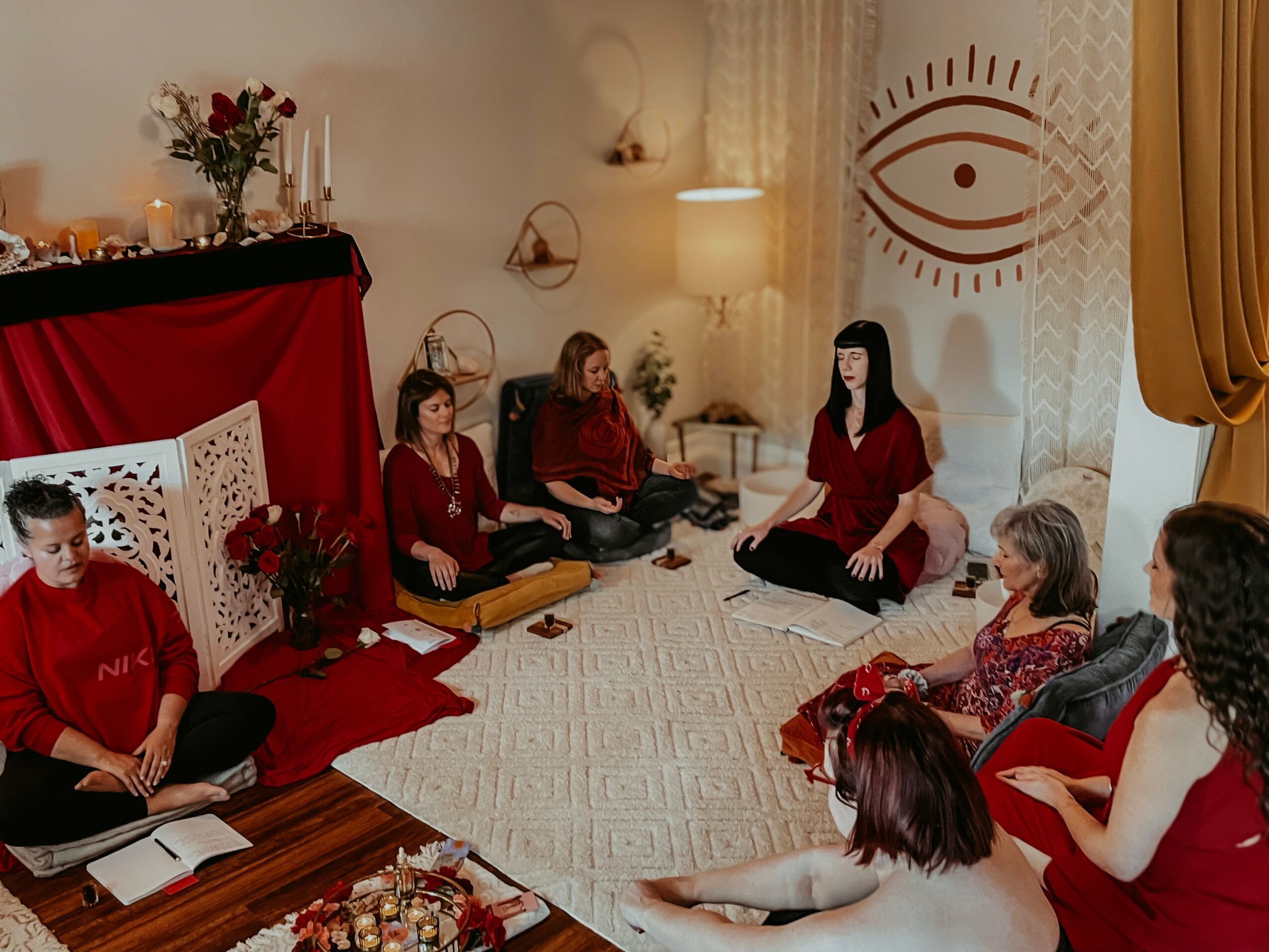 Group of women sitting on a rug in a room with a large eye symbol on the wall, flowers, and books.