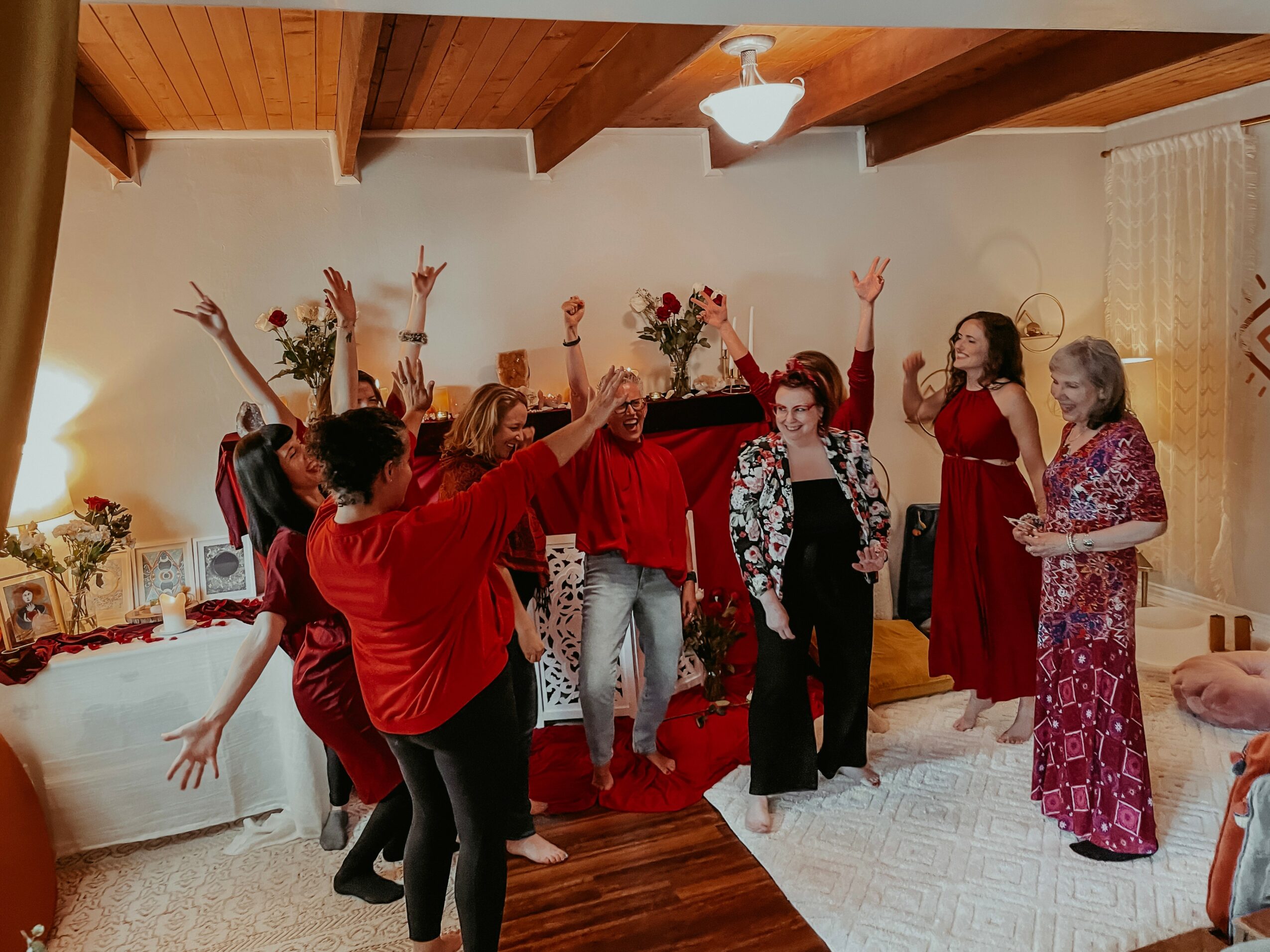 Group of women dancing and celebrating in a living room with wooden ceiling beams and a white rug.