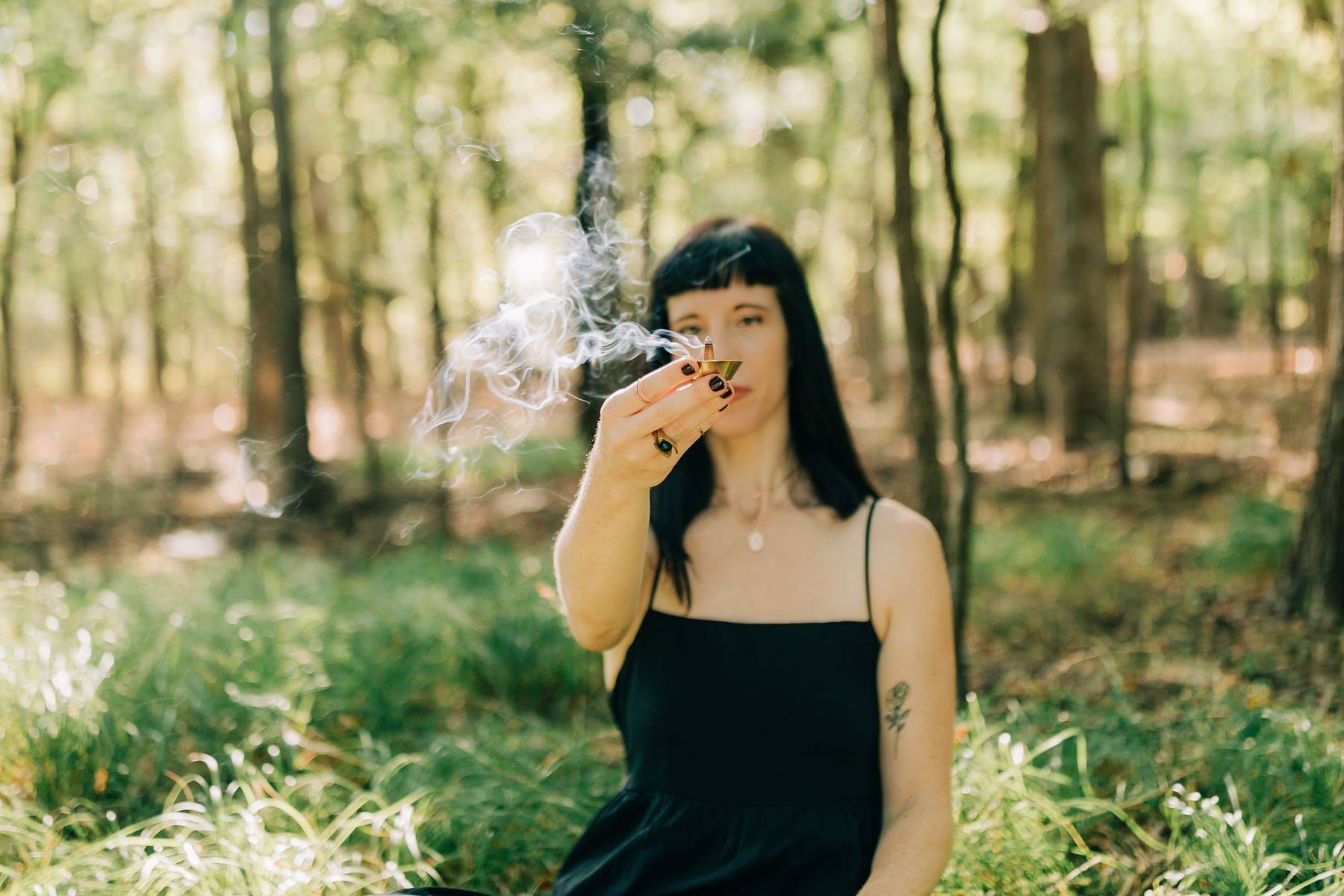 Woman with black hair and a tattoo on her arm smokes a cigarette in a forest.