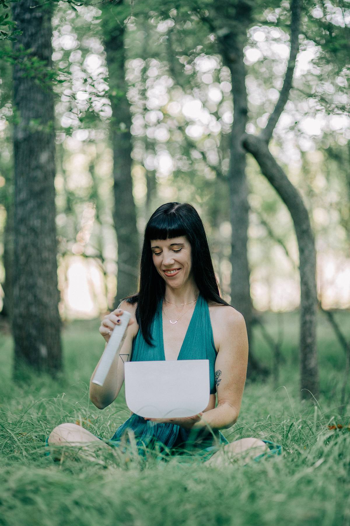 Young woman sitting cross-legged on grass in a forest, holding a bowl and a spoon, smiling.