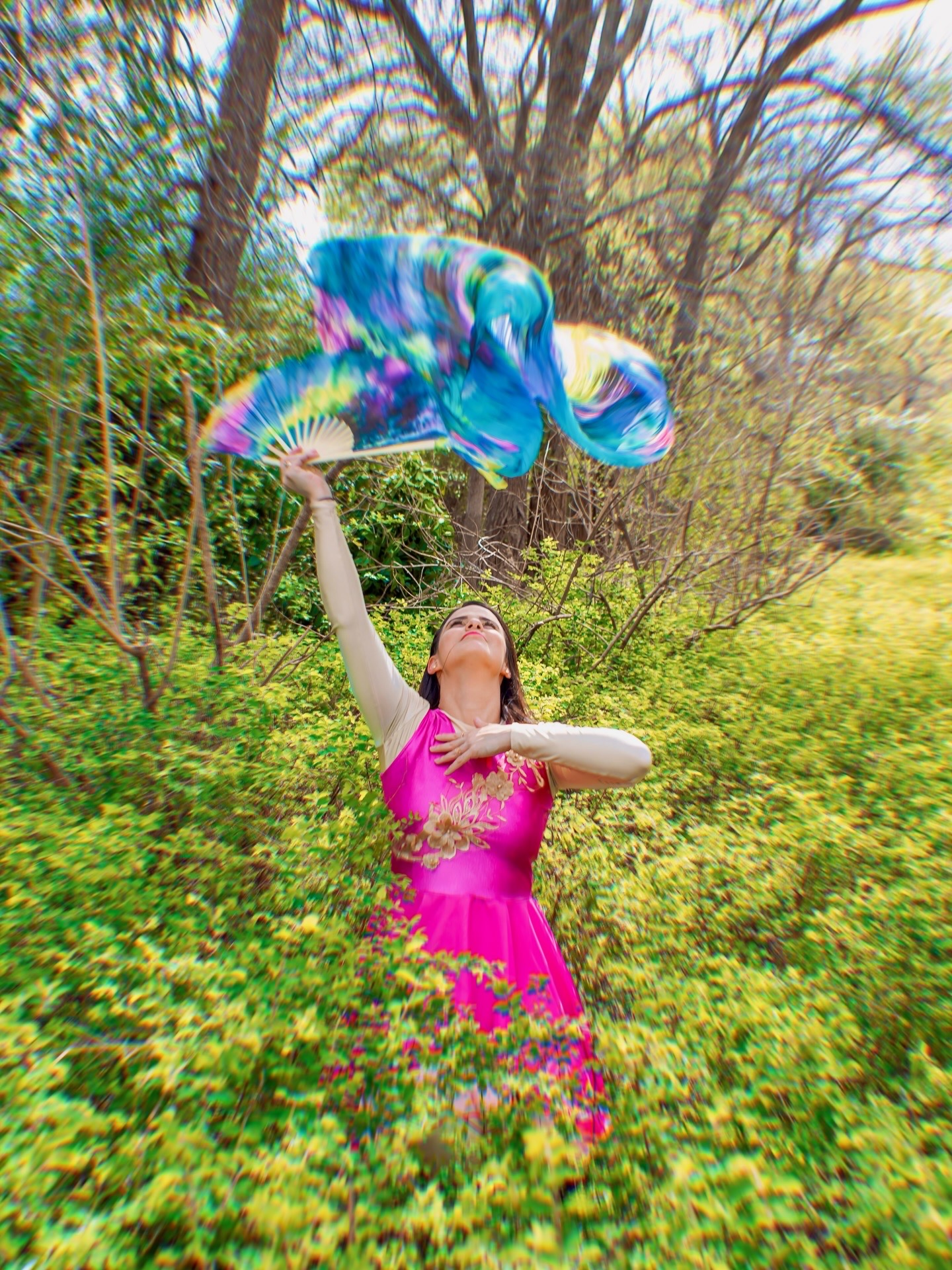 Woman in pink dress holding colorful balloon outdoors among green plants and trees.