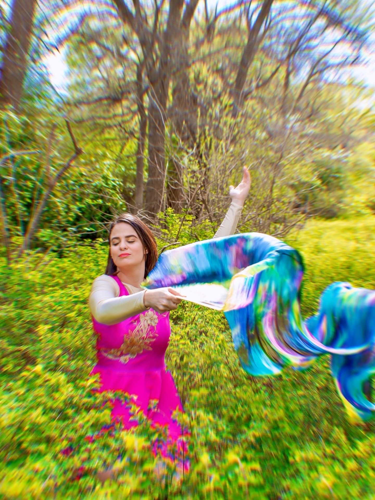 Woman in pink dress holding colorful fabric outdoors with trees and greenery in background.
