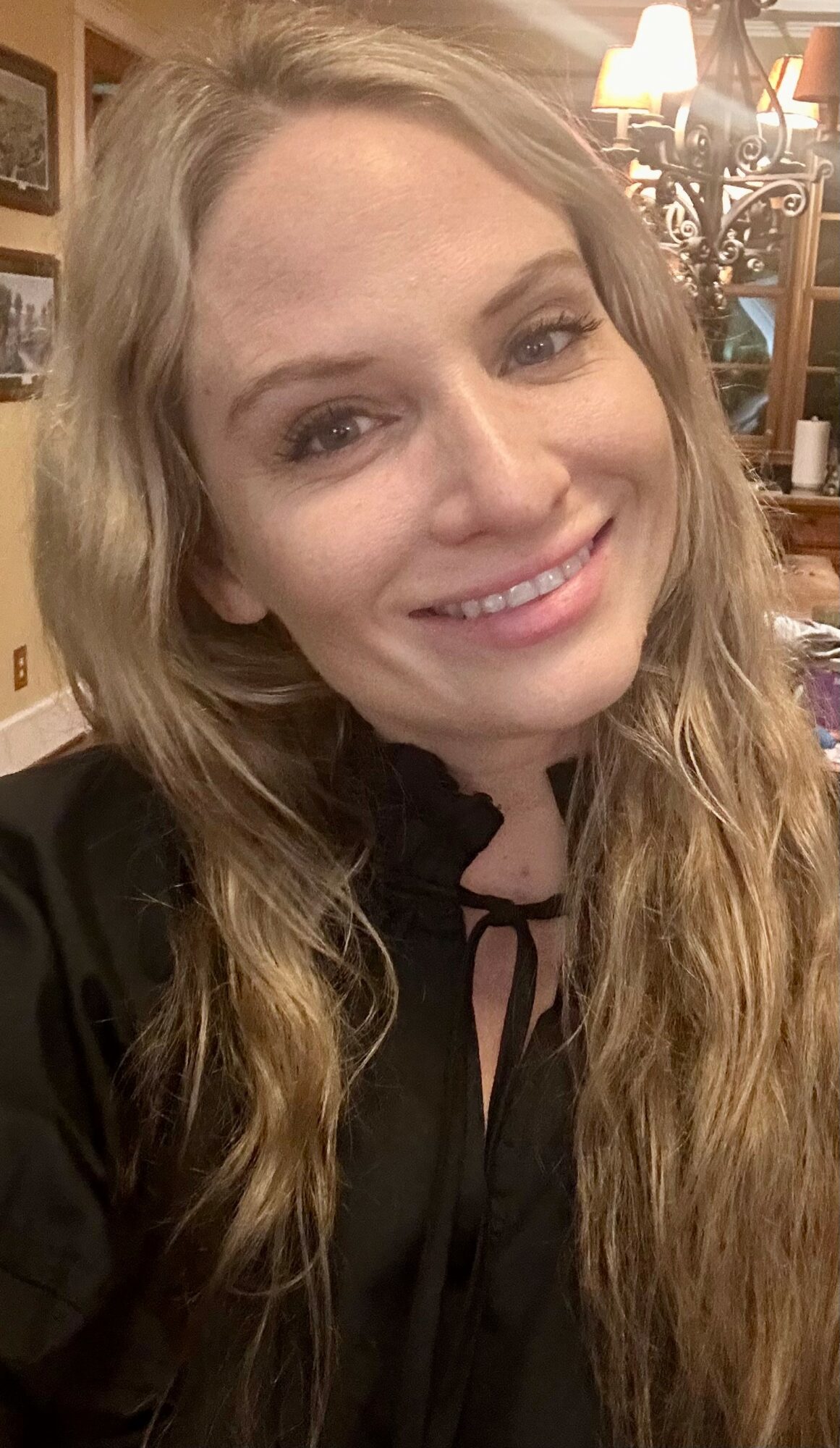 Young woman with long wavy hair smiling indoors, wearing a black top with a tie detail.