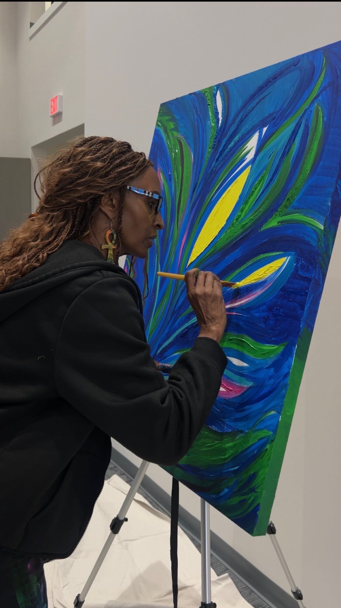 Person painting a colorful abstract on a canvas with a brush, standing indoors near a wall and easel.