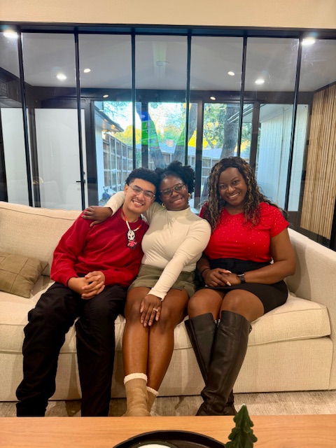 Three women sitting on a beige sofa, smiling, in a modern room with glass walls and wooden accents.