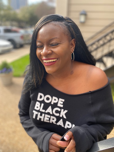 Smiling woman with long black hair, wearing hoop earrings and a black off-shoulder top with white text, outdoors.